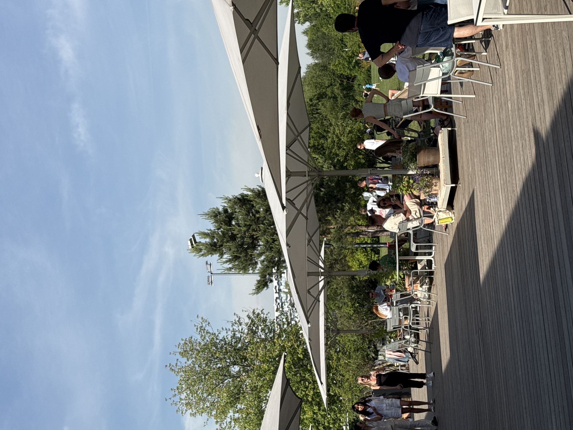 This image captures an outdoor cafe or restaurant terrace in Nemchinovo, Russia, on a sunny day. The foreground is dominated by a light-colored wooden deck, exhibiting distinct plank lines and shadows cast by various objects, indicative of strong sunlight from the right.
Several large, light gray octagonal sun umbrellas are strategically placed across the deck, providing shade for patrons. Beneath these umbrellas, numerous individuals are seated at white tables with matching chairs, engaging in conversations, dining, or relaxing. On the left side of the deck, three women are walking; one wears a light beige jumpsuit, another a black sleeveless dress, and the third a white two-piece ensemble.
In the mid-ground, a woman with red hair is seated at a table, facing left, while a man in sunglasses sits opposite her. Another woman in a light crop top and skirt stands facing away from the camera. Potted plants are dispersed across the deck, adding greenery to the seating areas, and a low wooden daybed-like structure with cushions and more potted plants is visible.
To the right, a man with dark hair, wearing a black t-shirt and denim shorts, stands with his back to the camera, hands on his hips. Behind the deck, a dense line of green trees and bushes forms a natural backdrop. Beyond these trees, a large grassy area is visible, where several people, including children in light-colored clothing, are present. In the far background, a modern building with a geometric, patterned facade can be seen through the trees, alongside tall poles topped with multiple floodlight fixtures. The sky above is a clear, light blue with faint, wispy clouds.