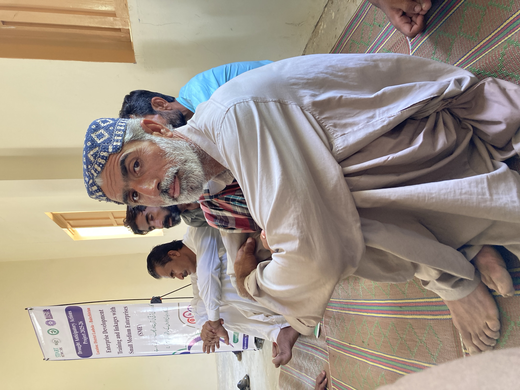 The image shows a group of men sitting on a woven mat in what appears to be an indoor setting in Goth Ali Marād, Pakistan. One man, closest to the camera, is wearing a patterned blue cap and a light-colored shirt. He has a grey beard and is looking directly at the camera. Several other men are seated behind him, some wearing traditional clothing such as white shirts and shawls. A banner is visible on the left side of the frame, with text written in English and possibly Urdu or another regional language. The banner mentions "Drought Anticipatory Actions Project 2025-26", and lists "District Lasbela - Balochistan" as a location. The banner also refers to "Enterprise Development Training and linkages with Small Medium Enterprises (SME)". The room has light-colored walls, a visible window frame near the ceiling, and the men are seated on a mat with multi-colored stripes. The scene suggests a workshop or meeting focused on enterprise development. The men are barefoot.