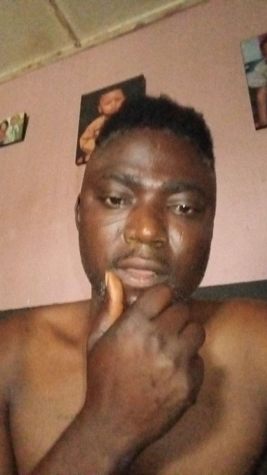 A shirtless man with dark skin and short, dark hair is looking directly at the camera, his hand resting on his chin. Three pictures are hanging on the wall behind him. The location, Sabon Tasha, Nigeria, is based on metadata.