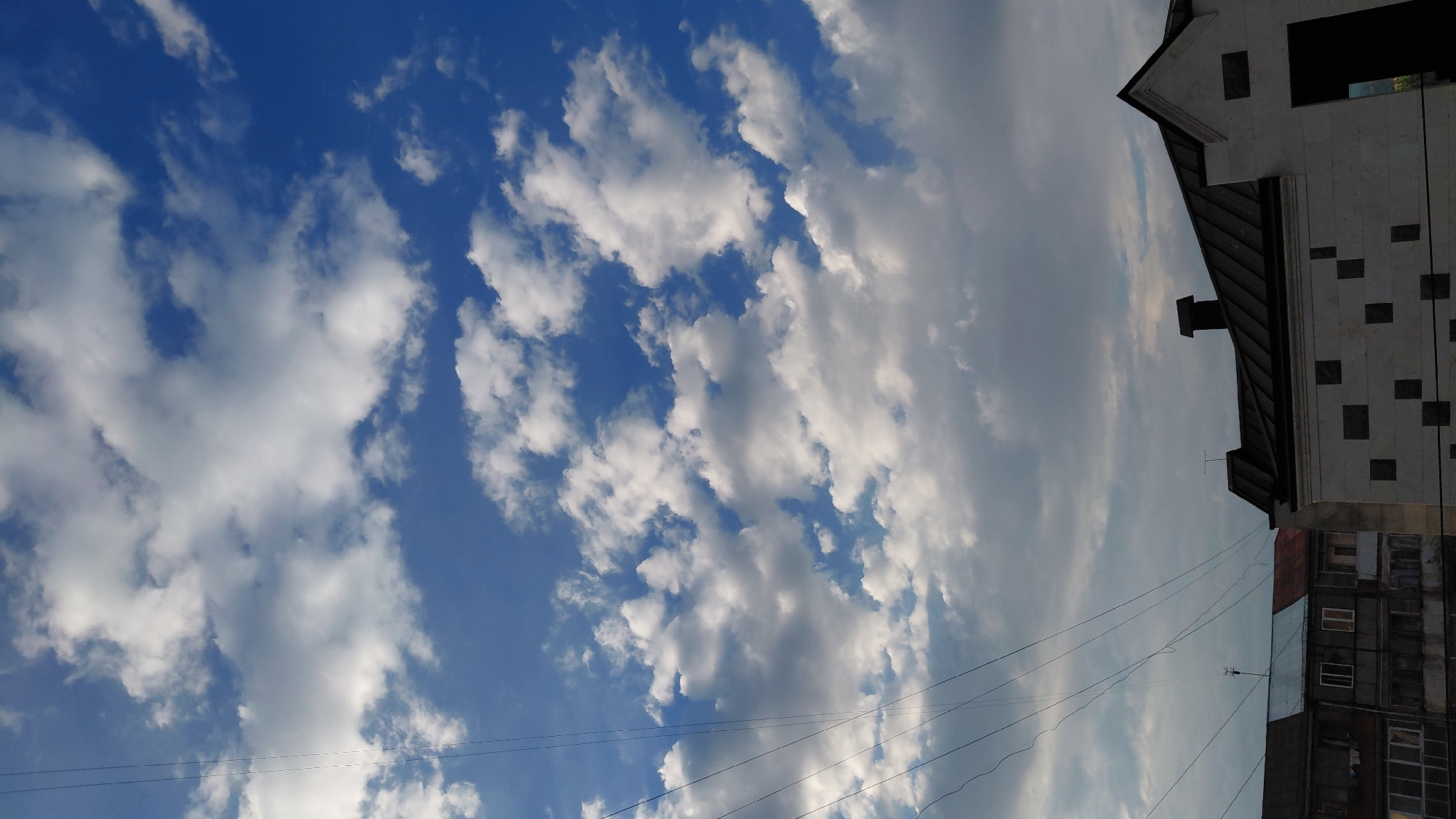 The image shows a view of the sky above buildings in Abovyan, Armenia. The sky is blue with scattered white clouds. Power lines are visible stretching across the upper left portion of the frame. In the foreground, the roofs of several buildings are visible, along with portions of their facades. One building in the foreground appears to be light-colored with dark square decorative elements. Another building to the left has a multi-colored roof and multiple windows. The overall impression is of a partly cloudy day over an urban or semi-urban setting.