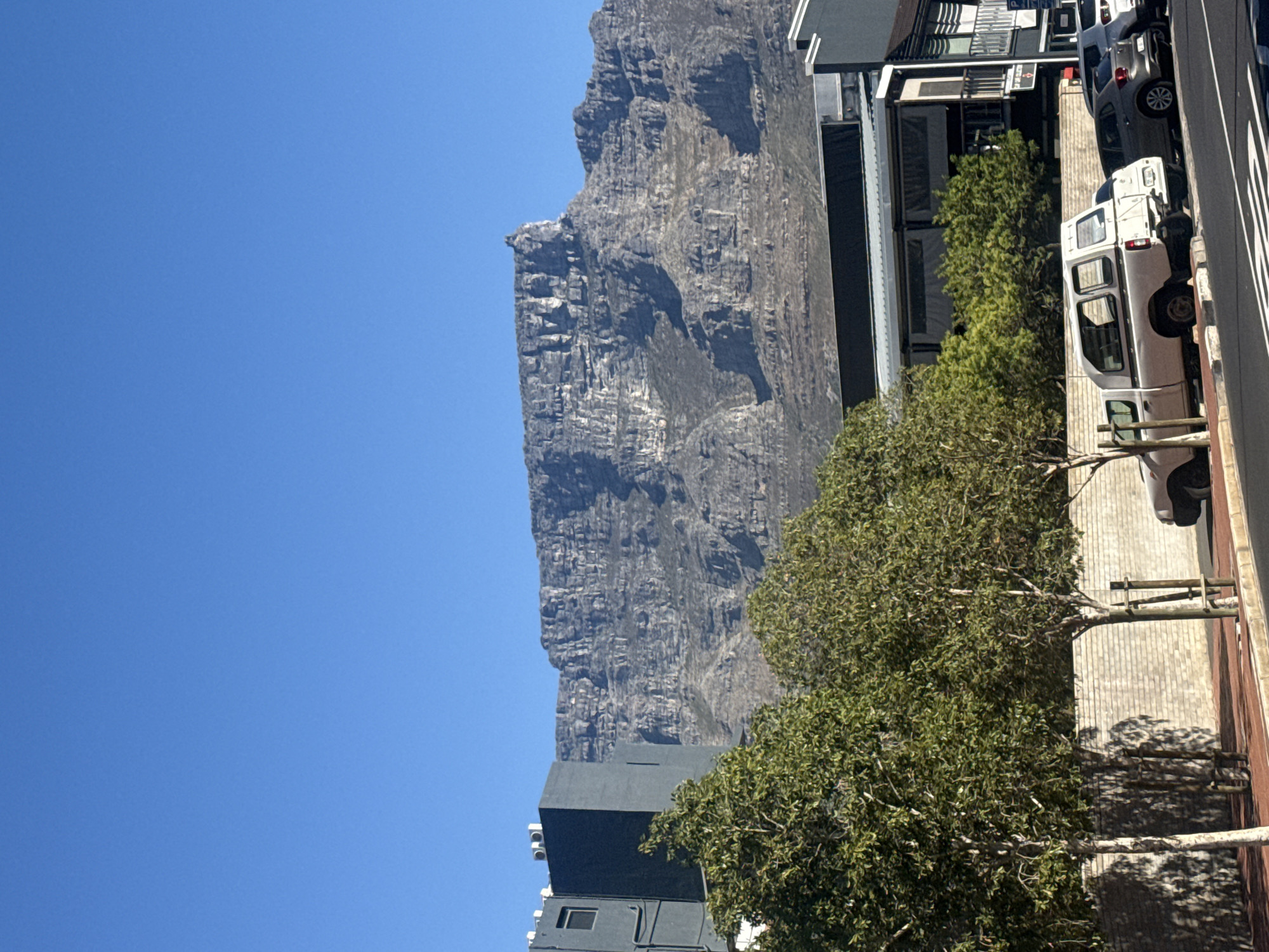 The image shows a view in Cape Town, South Africa, with Table Mountain dominating the landscape under a clear blue sky. A street scene is visible with parked cars, including a white van. Trees line the street, casting shadows on the pavement. Buildings are situated along the street, including a multi-story structure. The overall impression is a typical urban environment set against the backdrop of a prominent natural landmark.