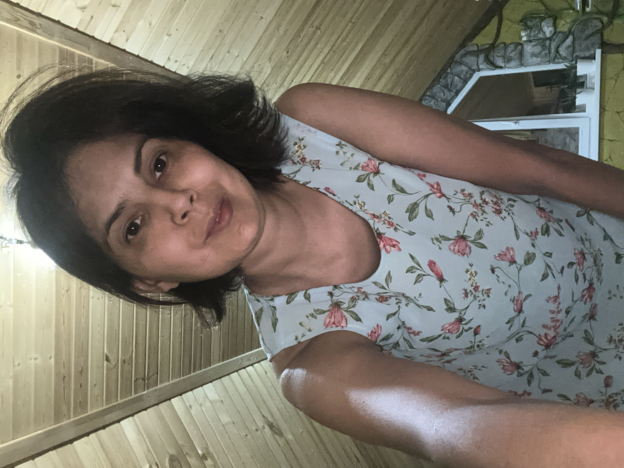 An indoor, medium shot photograph captures a woman from the chest up, with her head slightly tilted and facing the camera. She has dark, shoulder-length hair and is wearing a light blue, sleeveless dress adorned with a floral pattern of pink flowers and green leaves. Her right arm is extended forward, towards the camera, with her skin appearing slightly shiny. The background consists of a sloped wooden ceiling and a window with a stone-like frame. The overall lighting is warm, highlighting the woman's features and the textures of the environment.