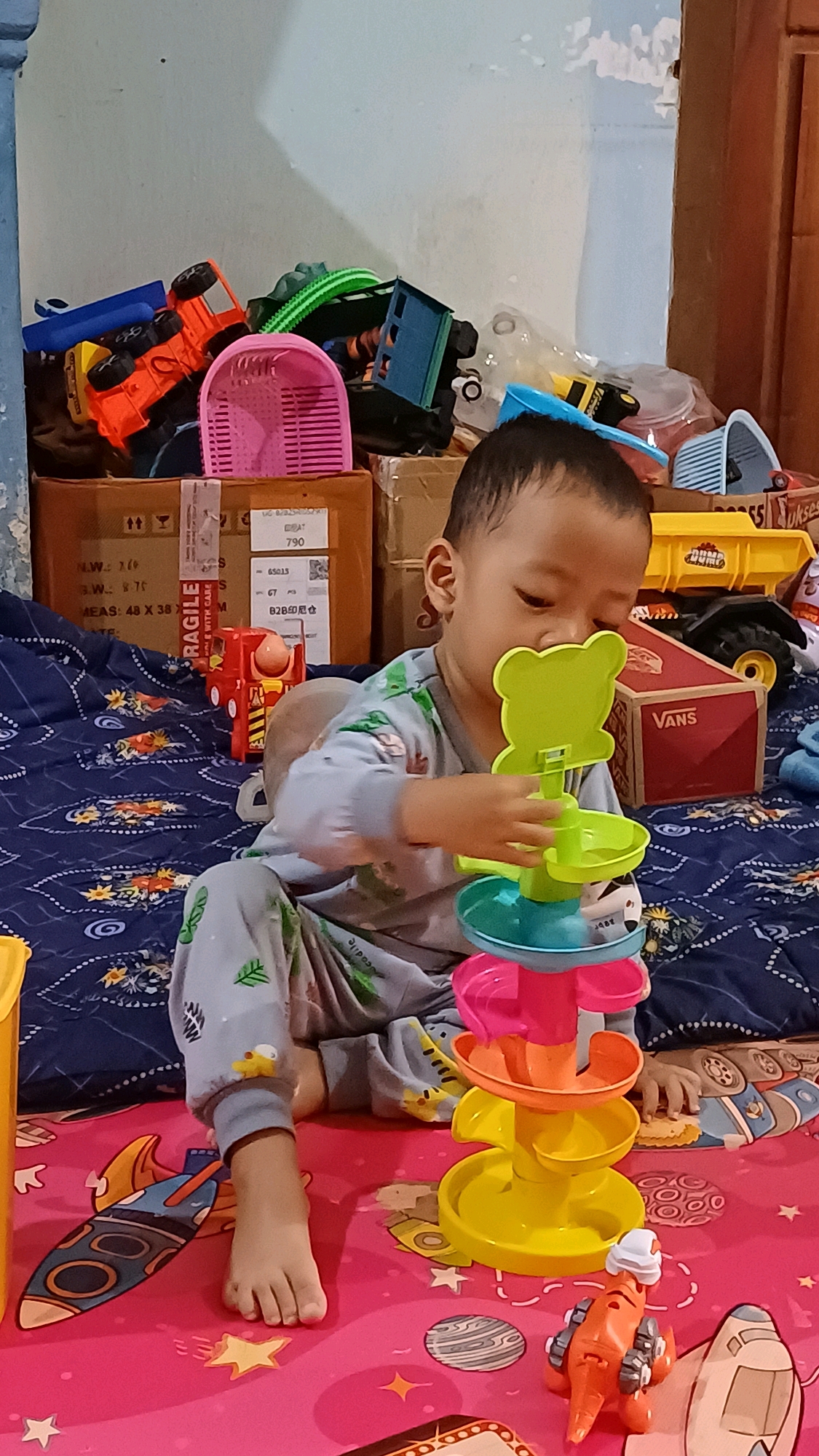 A young boy sits on a blue blanket spread over a pink mat, playing with a colorful toy tower. He is wearing grey pajamas with a yellow duck and other small yellow figures. The toy tower, which appears to be a ball drop toy, has multiple levels of colorful ramps and a green bear-shaped topper. The boy is reaching out with his right hand, interacting with the toy.
In the background, there are several stacked cardboard boxes and various toys, including a red and yellow toy truck with a cement mixer, a blue truck, a green ring, and other plastic toys. A red box with "VANS" logo is also visible. The environment suggests a child's play area, likely within a home. The lighting is indoor, and the scene appears to be taking place during the day. The presence of a blue pillar on the left side indicates a structural element of the room. The overall impression is one of a child engaged in play amidst a collection of toys and household items.