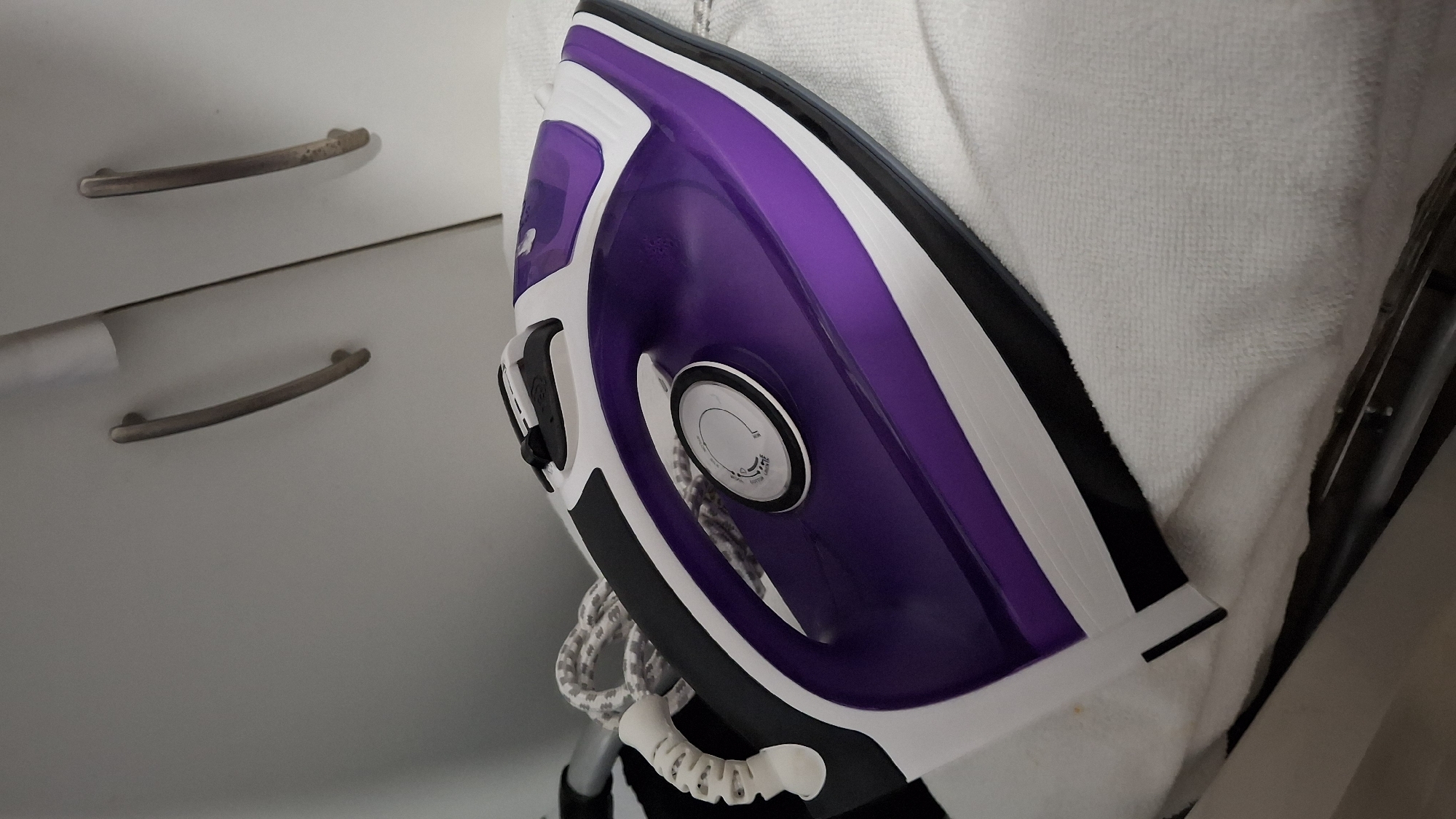 An iron with a purple, black, and white exterior rests on a white terrycloth towel. The iron is angled, with its handle in the foreground and its pointed tip extending away from the viewer. A coiled white and gray cord is visible behind the handle. The iron is positioned on a flat surface, likely a countertop or a table. In the background, a white cabinet with two curved silver handles is visible. The scene is illuminated by ambient light, casting soft shadows. This image was taken in a domestic setting.