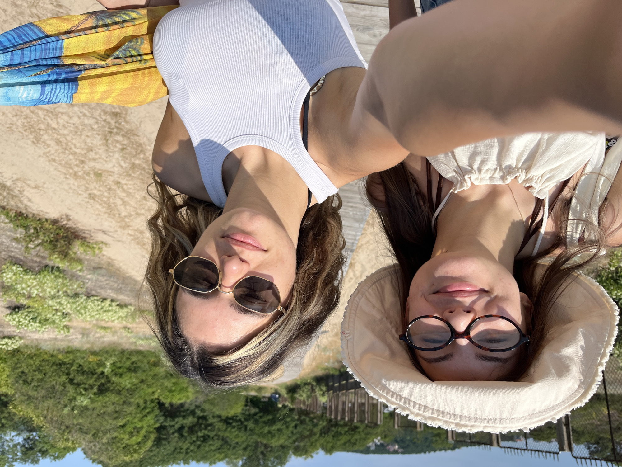 Two young women are taking a selfie on a wooden boardwalk. The woman on the left wears a wide-brimmed white hat and glasses. She has brown hair and is smiling. The woman on the right wears sunglasses and has blonde-streaked brown hair. She is also smiling. Both women are wearing tank tops. The woman on the right is holding a colorful towel. The background shows a sandy area with sparse vegetation and trees, with a fence and some buildings visible in the distance. The sky is clear and blue. The scene is likely located in Primorsko, Bulgaria, given the coastal vegetation and the presence of a boardwalk.