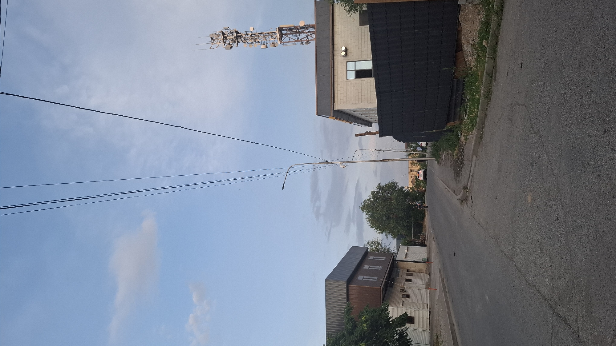 This image depicts a street scene in Shymkent, Kazakhstan, likely during daylight hours given the brightness. In the foreground, a paved road runs from the bottom left towards the background. On the left side of the road, there is a two-story building with a brown upper section and a white lower section. Above this building, a tree with green foliage is visible. Further down the street, a tall communication tower with multiple antennas stands on the right side of the frame. Adjacent to the tower, there is a light-colored building with windows. A dark, textured fence or wall runs along the property line to the right of this building. Overhead, multiple power lines traverse the sky. In the distance, several vehicles and what appear to be people are present on the road, suggesting a moderately active urban environment. The sky is a pale blue with scattered white clouds.