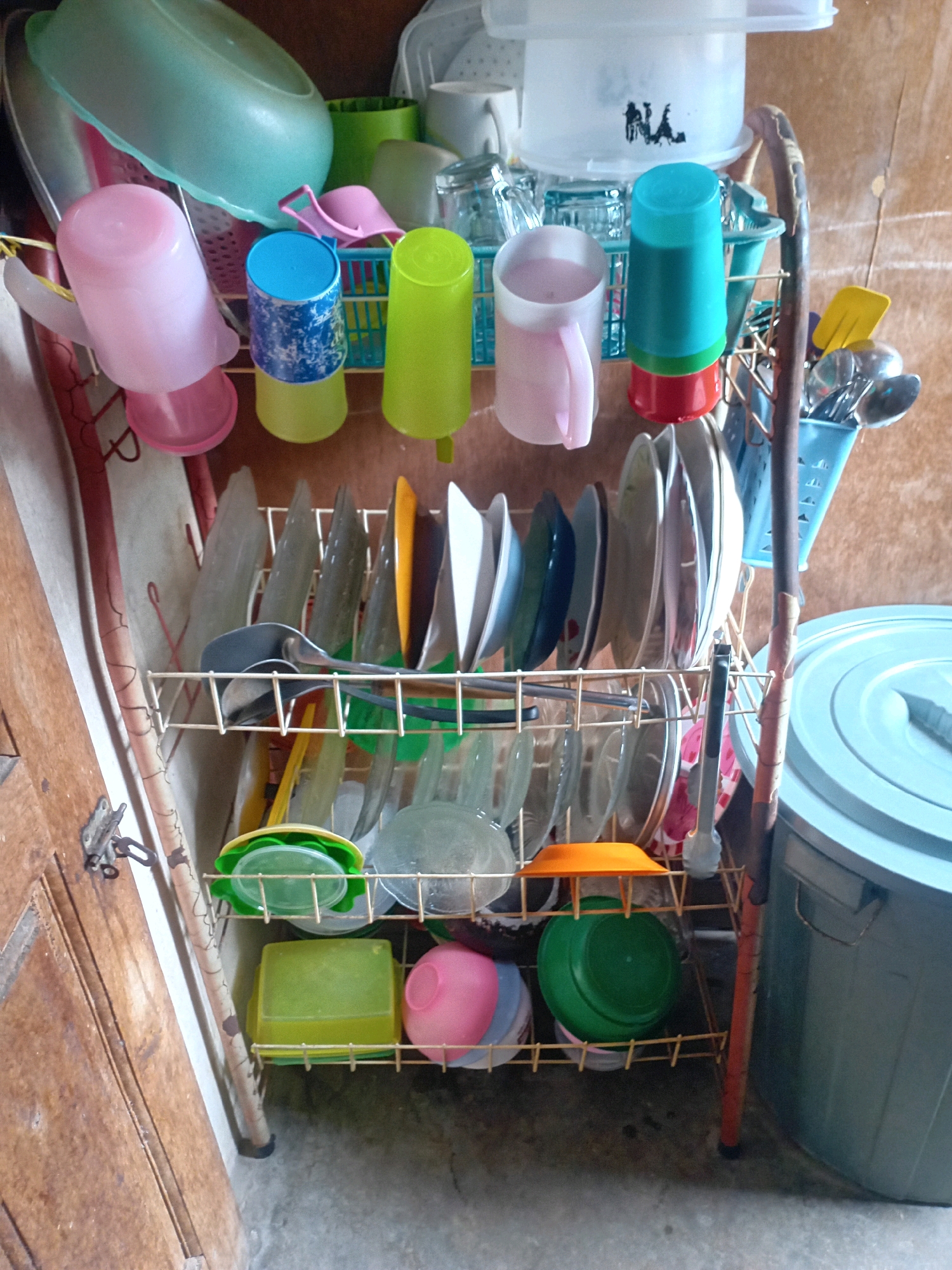 This image depicts a dish drying rack filled with various kitchenware. The rack is positioned in what appears to be a domestic or semi-commercial setting, potentially a kitchen or outdoor washing area. The scene is indoors, with wooden walls in the background.
Prominently featured are several stacks of plates, bowls, cups, and utensils. On the upper shelves, there are green and blue plastic cups, a pink mug, and what appears to be a larger green bowl. The middle shelves hold a collection of plates, some with colorful patterns, and various utensils like ladles and spoons. On the lower shelves, more bowls and containers are visible, including green and pink plastic containers, and a large green bowl. To the right of the dish rack, a large, light blue cylindrical container is visible, likely a storage bin or water container.
The overall impression is one of a functional, albeit somewhat cluttered, space dedicated to dishwashing and storage. The image captures a typical scene of domestic life in Pekanbaru, Indonesia, where such dish racks are commonly used to air dry dishes. The lighting suggests it is daytime, with natural light filtering into the space. The floor appears to be concrete. There are no people visible in the image.