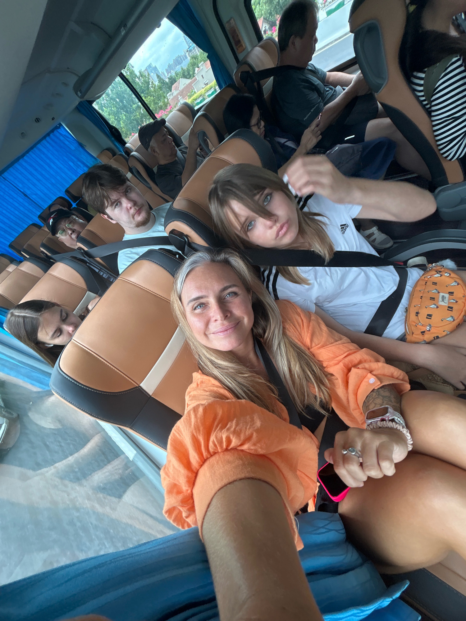 The image shows several people seated inside a bus, with a focus on a woman in the foreground taking a selfie. She is wearing an orange top and has a watch on her left wrist and a ring on her right ring finger. Beside her, a young woman in a white Adidas t-shirt is visible. Further back, other passengers are seated, some looking forward or at their phones. Through the windows, a cityscape with buildings and trees can be seen, suggesting an urban environment. The seats are upholstered in brown and black. The overall setting is consistent with being inside a tour bus or public transport. The specific location of Jing'an District, China cannot be determined from the visual information in the image alone.