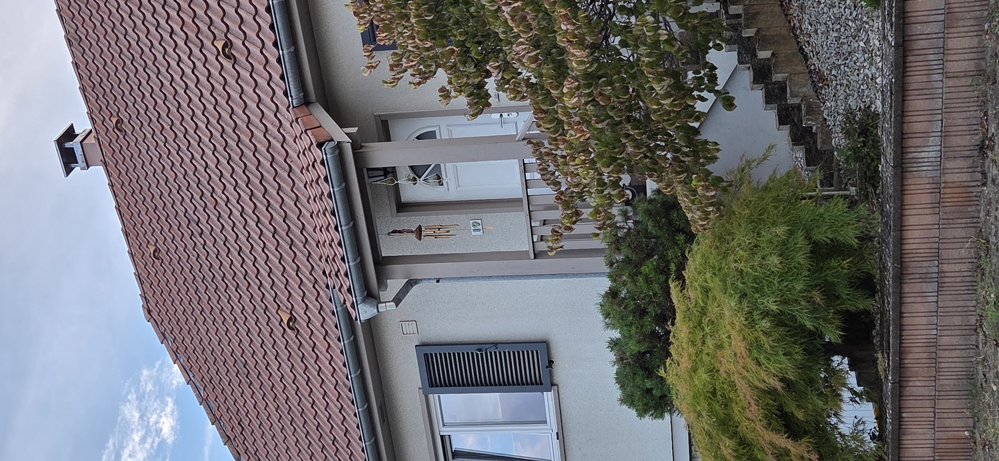 The image shows the front of a residential house in Kilstett, France, on a cloudy day. The house has a red-tiled roof and a light-colored exterior wall. To the left of the entrance, there is a window with dark grey shutters. The entrance features a white door, a porch with a railing, and a number "19" displayed on the wall. A wind chime hangs near the entrance. Two trees are visible in front of the house: a large, leafy green tree to the right of the entrance, and a more delicate, feathery green tree to the left. There is a set of concrete stairs leading up to the entrance, flanked by gravel and greenery. A low brick wall with vertical grooves runs along the bottom of the image, separating the property from the foreground. A chimney with a metal cap is visible on the roof. The sky is partly cloudy.