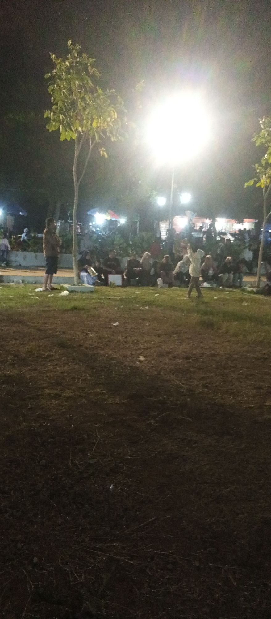 The image depicts an outdoor scene at night, likely in Cermo, Indonesia, illuminated by a bright overhead light. In the foreground, a grassy area is visible, with scattered white debris. In the midground, a man stands alone on a paved area, facing towards the right of the frame. Behind him, a group of people are seated on the ground. Further in the background, there is a gathering of people, with some under umbrellas or awnings, suggesting a market or event. Two trees are present, one in the left midground and another on the far right edge of the frame, their leaves illuminated by the artificial light. The overall atmosphere appears to be casual and social.