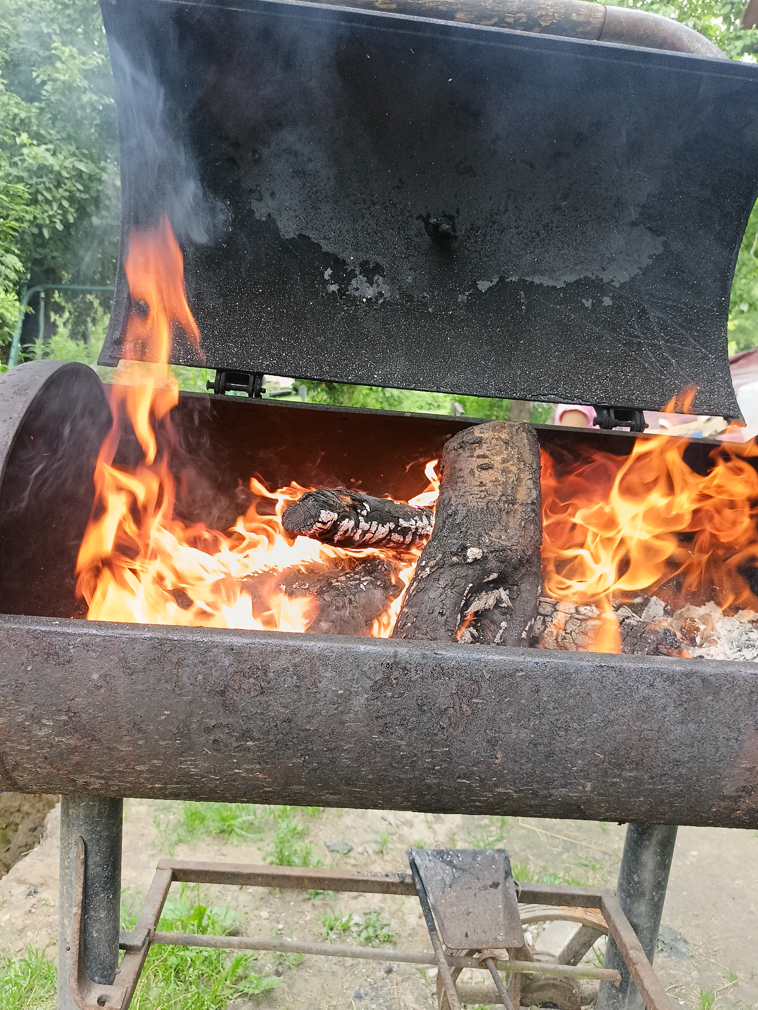 The image depicts a close-up view of a smoking barbecue grill with a fire burning inside. The grill is made of dark, weathered metal and is supported by metal legs. Flames are visible within the grill, along with burning logs. Smoke billows upwards from the grill, obscuring some of the background. The environment appears to be outdoors, with greenery and trees visible in the background. There are hints of people in the far background, suggesting it is a social gathering. The location is identified as Lagovskoe, Russia.