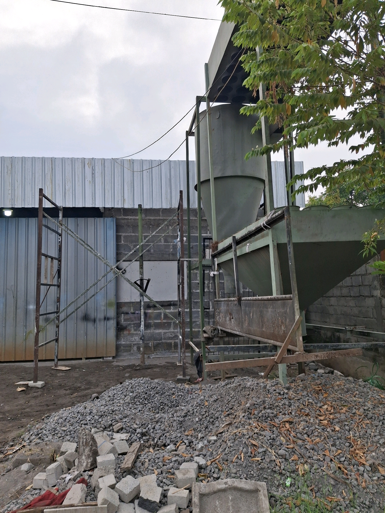 The image depicts an outdoor industrial area, possibly a construction or material processing site, located in Lumajang, Indonesia. In the foreground, a large pile of gravel and broken concrete blocks lies on the ground, with some scattered leaves. A few pieces of red fabric are also visible among the debris. Behind the gravel pile, a section of a metal structure with diagonal bracing stands, part of a larger industrial apparatus. This apparatus includes large, green-painted hoppers and chutes, suggesting a system for handling bulk materials. The structure is supported by metal scaffolding. In the background, a corrugated metal building with a roller door and a visible light fixture forms a wall. Adjacent to this building is a wall constructed of dark cinder blocks. Lush green foliage from a tree hangs into the frame from the upper right. The sky above is overcast with clouds. The scene appears to be a functional, albeit somewhat unkempt, industrial workspace.