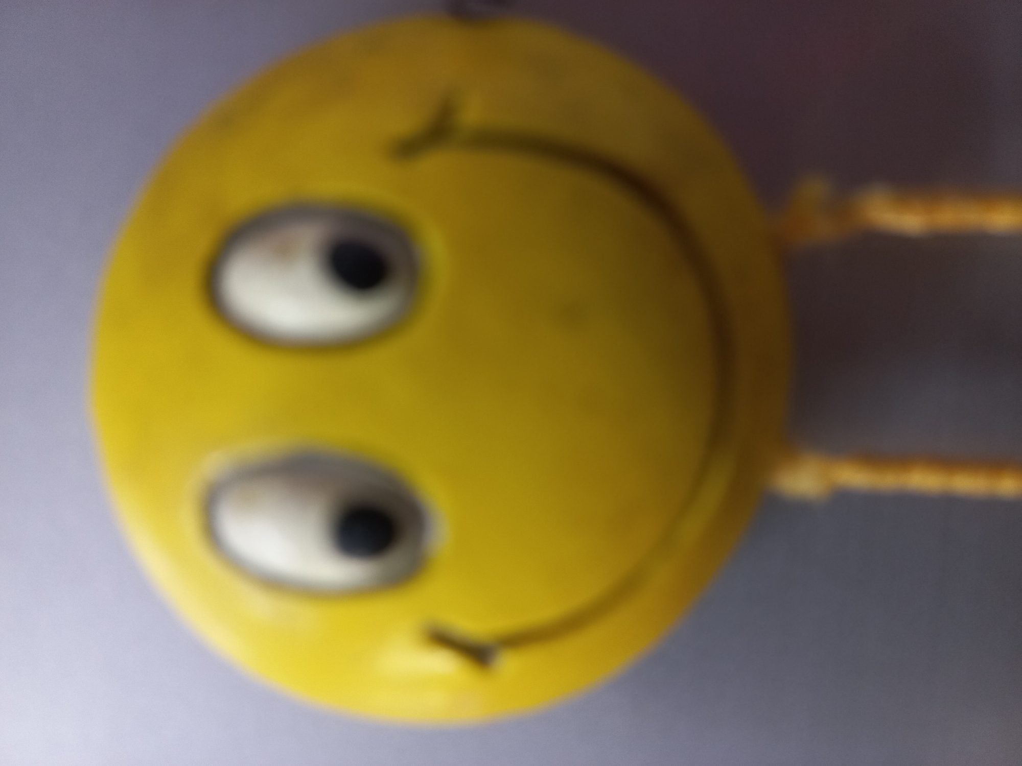 A blurry, close-up image shows a yellow, smiling toy figure with two large white eyes, each containing a black pupil. The figure has a wide, curved smile. Two thin, yellow, string-like legs hang down from the bottom of the figure. The background is a muted, out-of-focus gradient of light purple and grey. The toy appears to be made of plastic or a similar smooth material. The image lacks any identifiable context related to the city of Ashgabat, Turkmenistan.