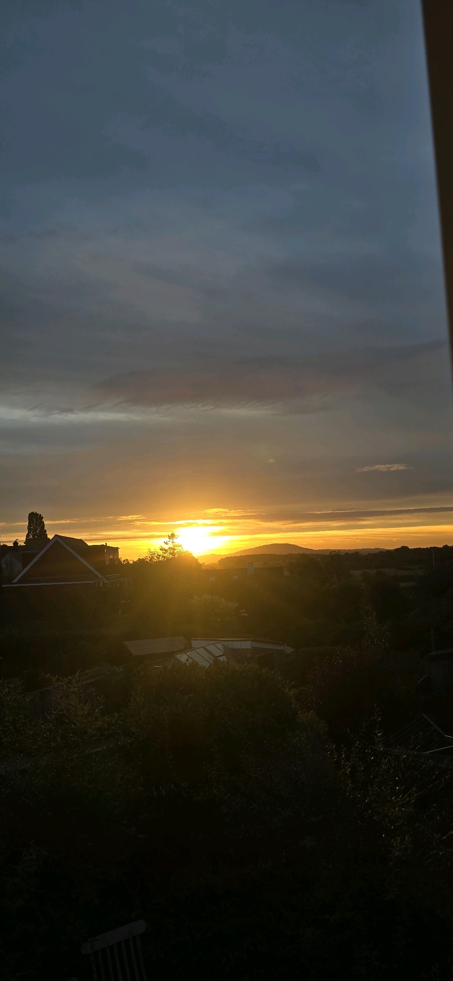 A serene sunset unfolds over the rural landscape of Bayston Hill, United Kingdom. The sun, a brilliant orb of golden light, dips below the horizon, casting a warm glow across the sky and silhouetting a distant hill. Wispy clouds, tinged with shades of orange and pink, drift across the dusky blue expanse. In the foreground, the dark silhouette of trees and shrubs frames the scene, with the roofline of a house and a greenhouse peeking through. The overall impression is one of tranquility and the quiet beauty of nature at dusk.