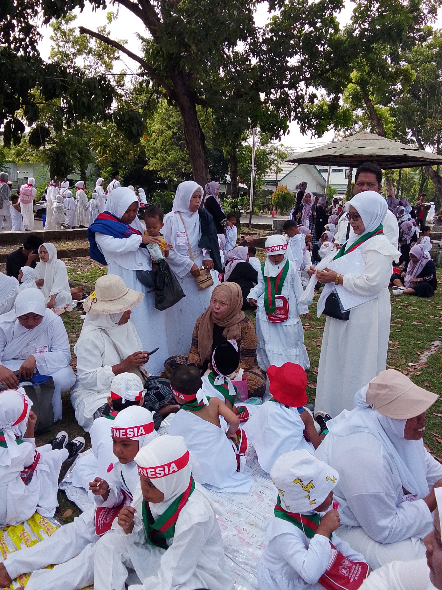 In Cirebon, Indonesia, a group of people, primarily children and adults, are gathered outdoors on a grassy area, possibly for an event or religious observance. Many individuals are wearing white attire, including headscarves and robes, suggesting a communal activity like a pilgrimage or a school event. Some children are wearing red and white headbands with "INDONESIA" printed on them, indicating a national or cultural theme. Several adults are supervising the children, with some interacting with them, such as one woman holding a baby and another assisting a child with clothing. Objects visible include bags, a stroller, and a water dispenser. The environment features trees, a shaded pavilion, and a white building in the background, suggesting a park or a public space within the city. The overall atmosphere appears to be one of togetherness and organized activity.