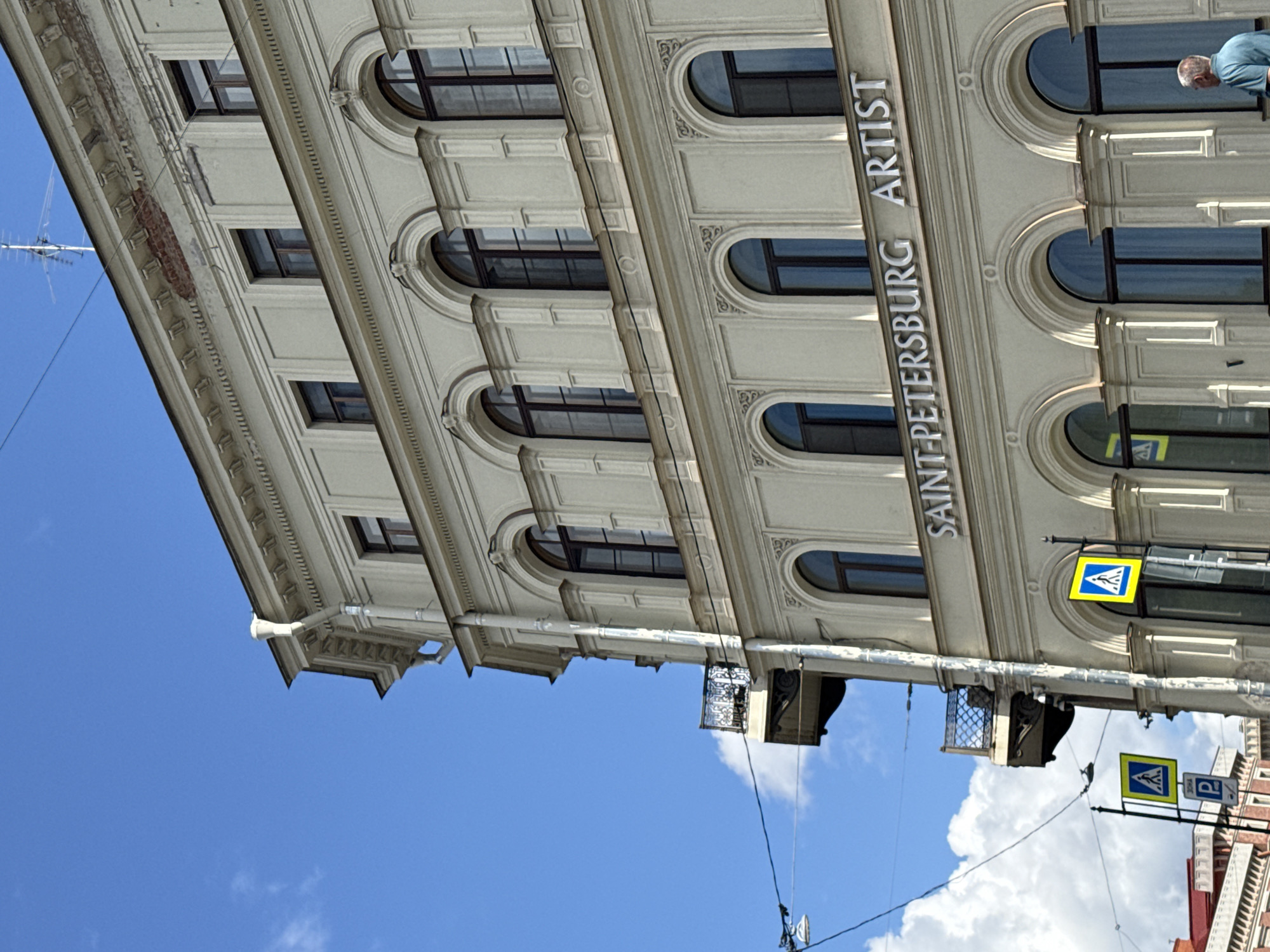 This image shows a street view of a building in Saint Petersburg, Russia, under a bright blue sky with scattered clouds. The building is light-colored with ornate architectural details, including arched windows, decorative moldings, and a prominent cornice. The words "SAINT-PETERSBURG ARTIST" are displayed in silver lettering on the facade. Several traffic signs are visible, including pedestrian crossing signs and a parking sign. There are also power lines and streetlights. In the lower right corner, a man in a blue shirt is partially visible, looking towards the building. The overall impression is of a historic urban environment bathed in sunlight.