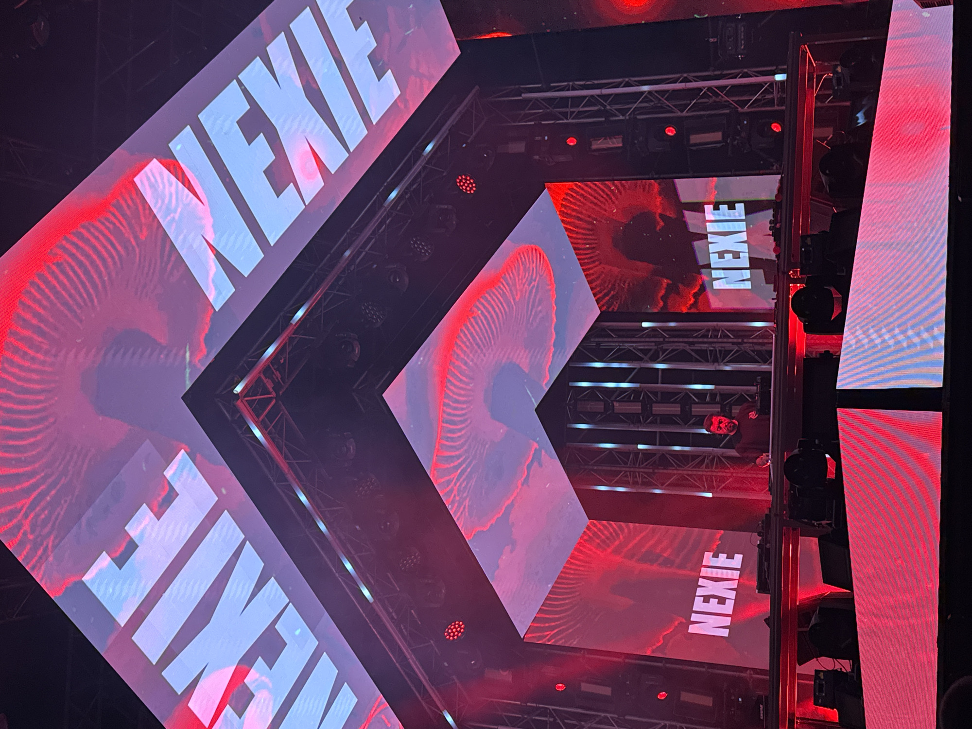 The image shows a concert stage with LED screens displaying the word "NEXIE" in large, white block letters. The screens also feature red, swirling graphical patterns. Stage lighting and rigging are visible, including a person on a higher platform. The location is a concert venue, which is likely in Budva, Montenegro, given the provided context.