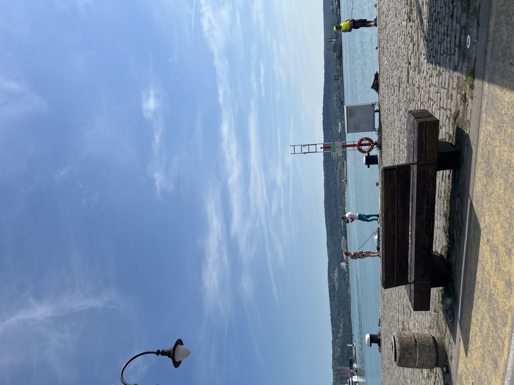 This image depicts a sunny daytime scene on a cobblestone pier or waterfront in Varna, Bulgaria, overlooking a calm body of water, likely Varna Bay. The sky is clear blue with sparse, wispy clouds.
In the immediate foreground, a dark wooden public bench with concrete supports is positioned centrally, facing the water. To its left, a cylindrical concrete waste bin is visible. The ground surface consists of rough cobblestones transitioning to a smoother, yellowish-grey concrete strip closer to the viewer. A partial view of an antique-style street lamp with a white globe fixture is present on the far left.
Three individuals are present on the pier. Towards the left-center, a woman wearing a light-colored top and patterned shorts stands near the water's edge, holding a fishing rod directed into the water. Beside her, a man in a white t-shirt and blue jeans is looking down at a smartphone in his hands. Further to the right, a solitary man wearing a bright yellow-green t-shirt or vest and a matching cap stands at the pier's edge, also holding a fishing rod and facing the water.
Between the bench and the solo fisherman, a red and white striped pole with a ladder-like attachment extends upwards. An orange lifebuoy hangs from this pole, and a small white rectangular sign with illegible text (possibly "112") is affixed below it. A grey utility box or cabinet is situated near the pole. Dark-colored mooring bollards are spaced along the pier's edge.
The background features a verdant, hilly coastline across the water, dotted with scattered white and light-colored buildings. A lighthouse or navigational beacon is visible on the left side of this distant shoreline. Several ships, including a white yacht on the far left and multiple larger vessels (possibly cargo or naval ships), are either docked or anchored in the bay. The overall scene suggests a leisurely, recreational atmosphere at a working port or harbor.