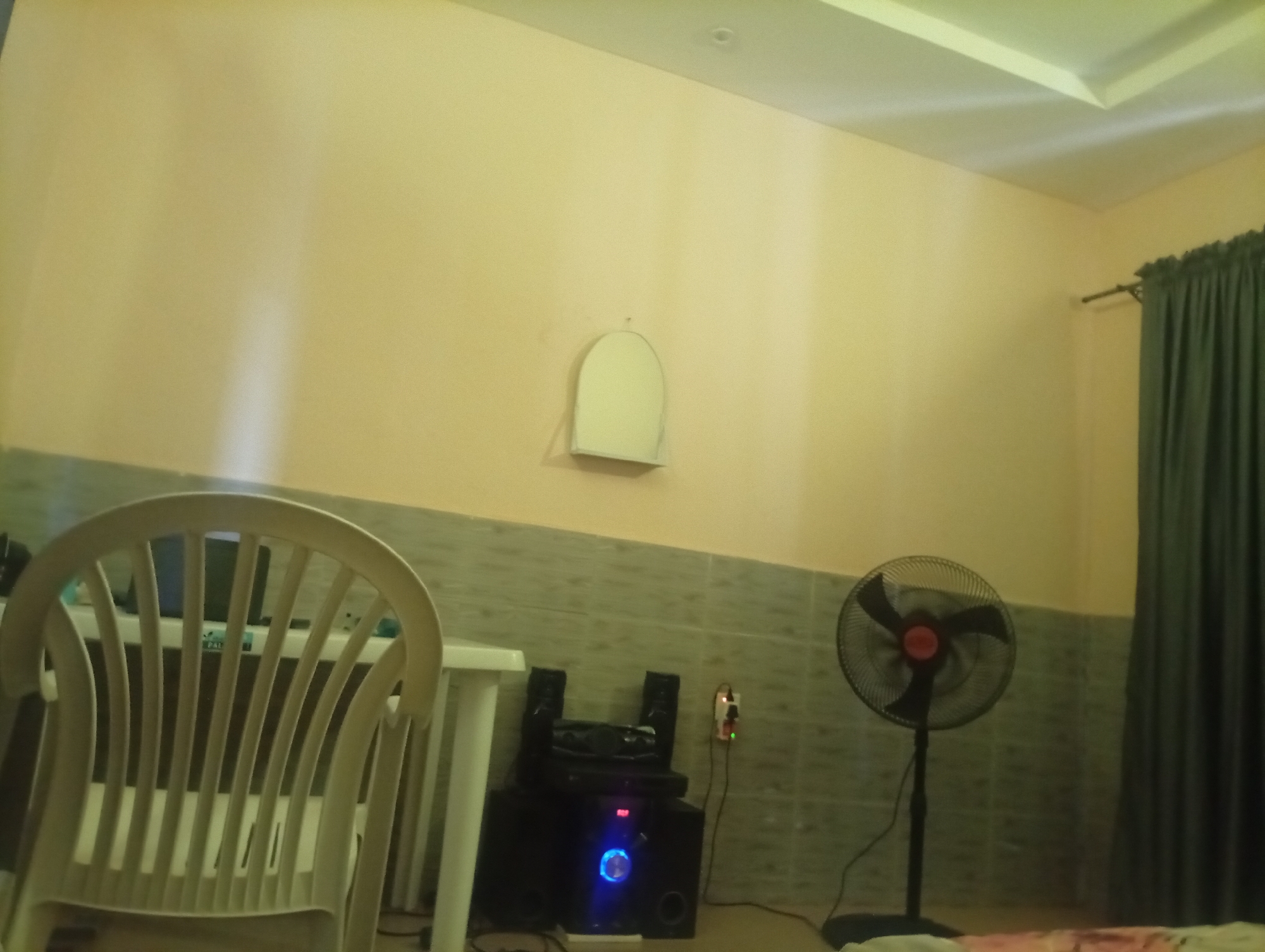 The image shows an indoor scene. In the foreground on the left is a white plastic chair with a curved back and slatted seat. To the right of the chair is a white table. Behind the table is a black speaker system with blue lighting on the front of one of the units. To the right of the speaker system is a black oscillating fan on a stand. In the background on the right is a dark green curtain hanging from a rod. The walls are a light yellow color, with a tiled pattern on the lower portion of the walls. A white, arched object is mounted on the wall above the speaker system. The ceiling is white and has recessed lighting. The floor is not clearly visible.