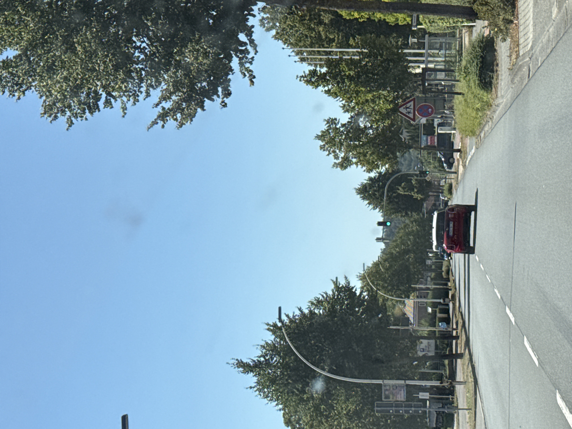 The image shows a street scene in Bad Lauterberg im Harz, Germany, on a clear day. A red car is driving on the road, with trees and buildings lining the street. There are traffic lights and street signs visible. A bus stop is also present on the right side of the road. The overall impression is of a suburban or town environment.