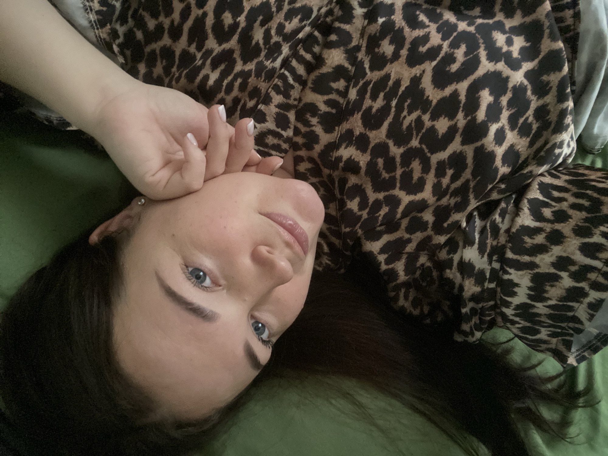 A woman with fair skin, blue eyes, and dark hair is lying down. Her right hand is near her face. She is wearing a leopard print top. The background appears to be a green surface. The location is not identifiable as being in Sochi, Russia, based on the visual information provided.