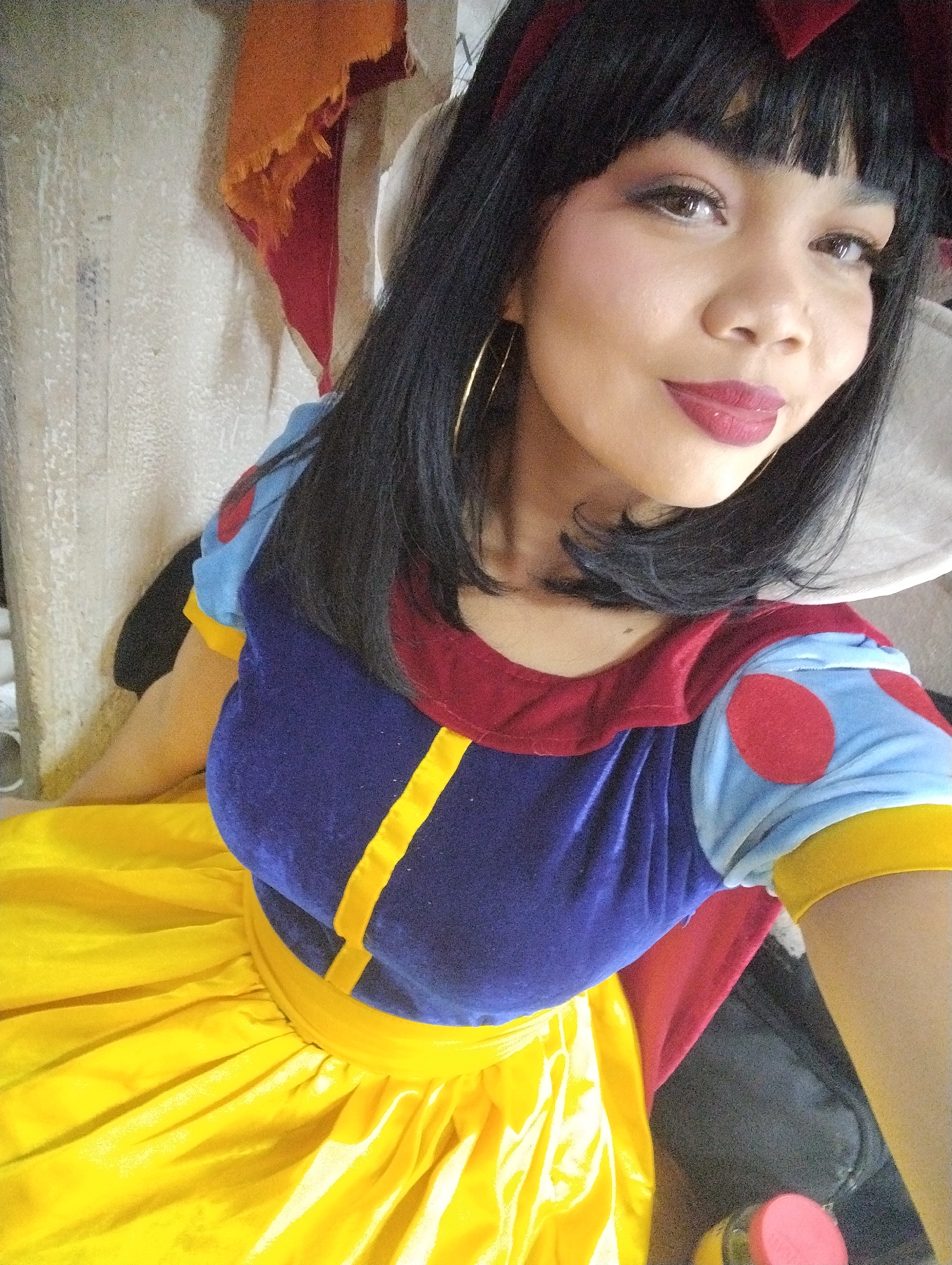 A woman is posing indoors while dressed as Snow White. She has black hair, dark red lipstick, and gold hoop earrings. Her dress is a combination of yellow, blue, and red. The environment is unclear, but there are fabrics and household objects in the background. There is no evidence to place the location in Porlamar, Venezuela.