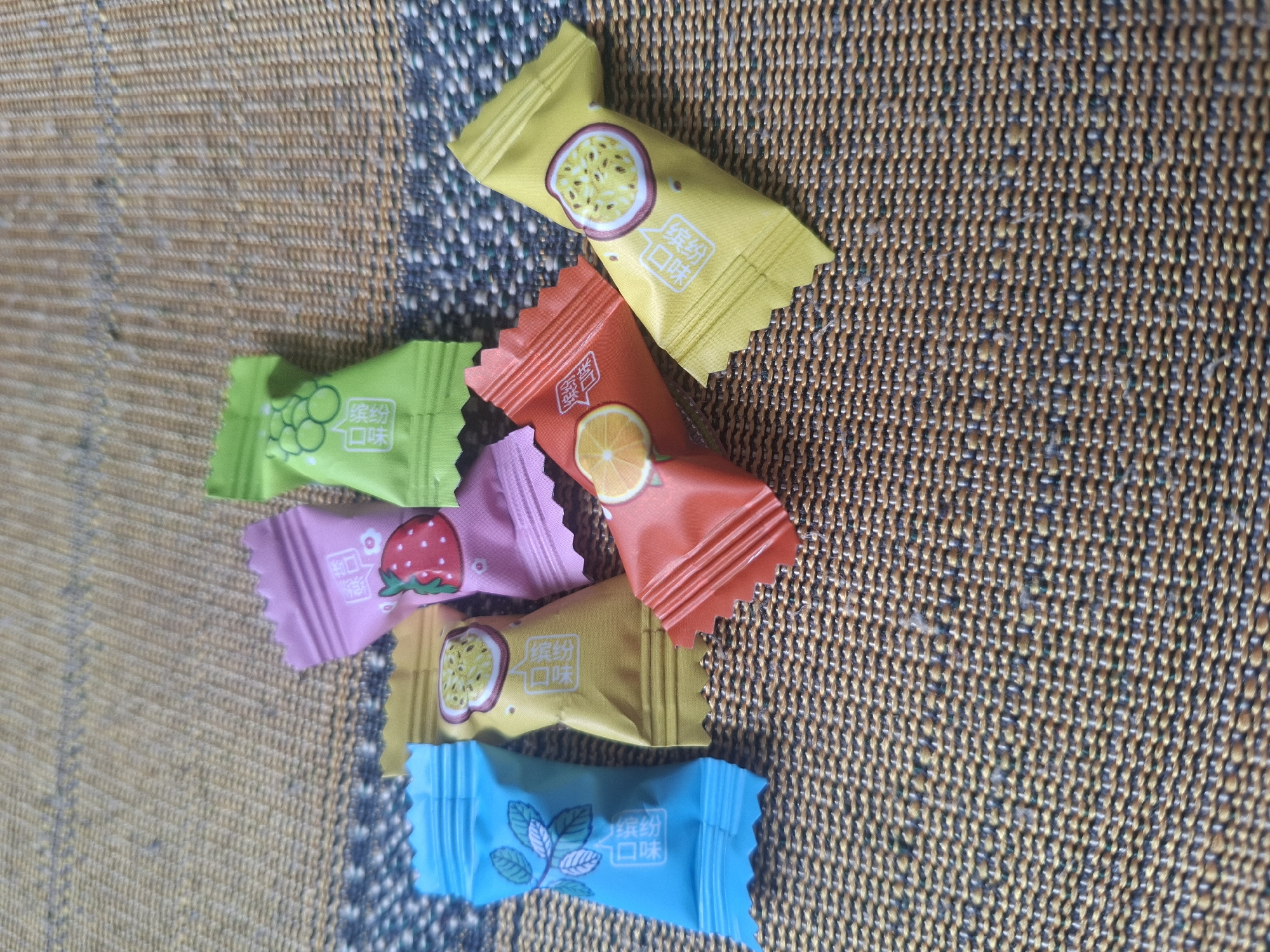 A pile of individually wrapped candies sits on a woven, textured surface, likely a placemat or tablecloth. The candies are in various bright colors: yellow, green, pink, red, and blue. Each wrapper features a simple illustration and Chinese text.
The yellow wrappers depict a slice of passionfruit. The green wrapper shows a cluster of grapes. The pink wrapper has a drawing of a strawberry. The red wrapper displays a slice of orange. The blue wrapper features a sprig of mint leaves. The Chinese text on each wrapper appears to be the same, likely indicating "flavor."
The lighting suggests daytime, possibly indoors. The overall impression is a colorful collection of sweet treats. No people are visible, and the activity is simply the presentation of the candies. The location context of ДТС «Чишки», Russia, is noted, but no specific visual cues in the image itself directly indicate this location.