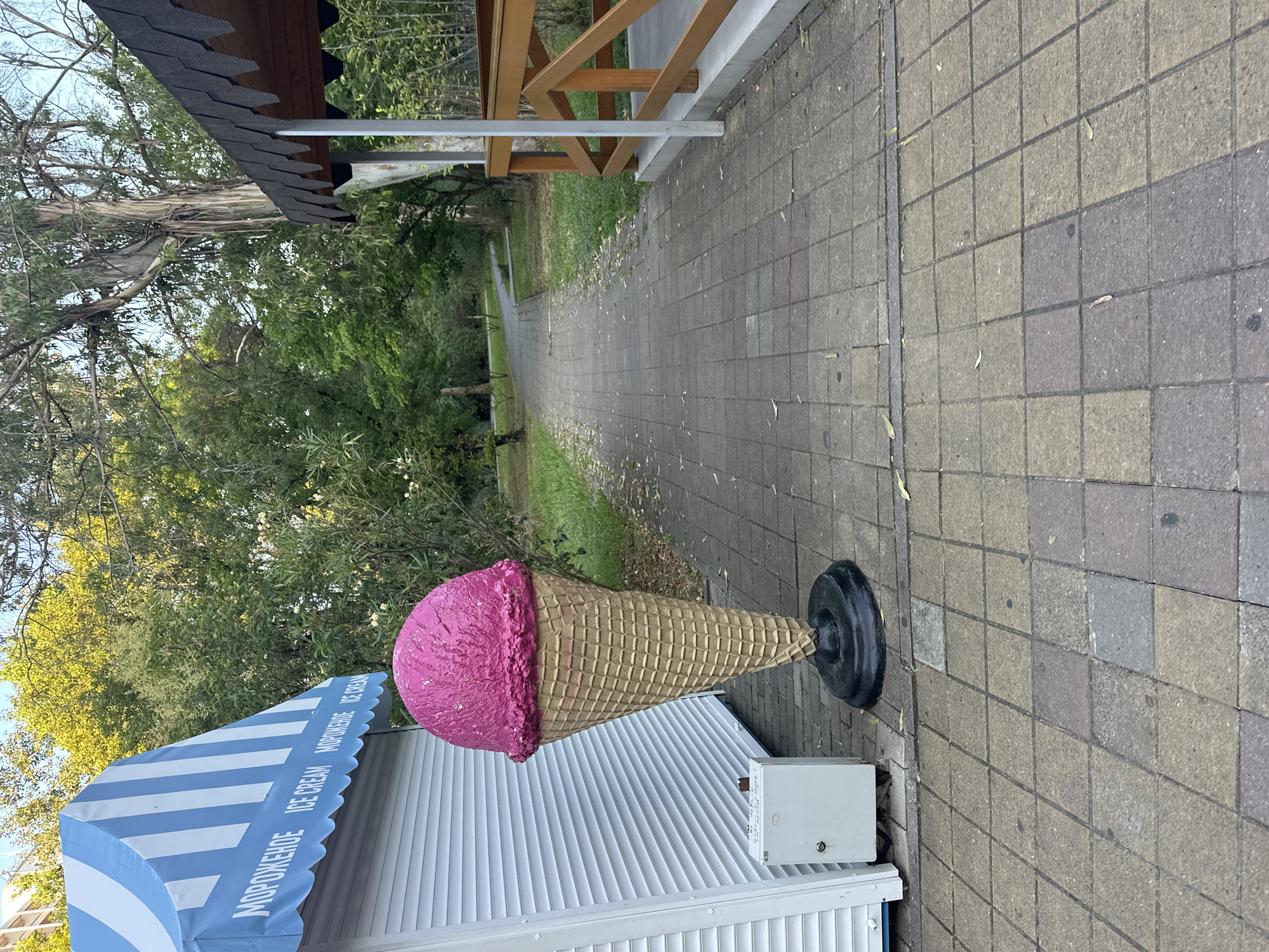 The image depicts an outdoor scene on a paved pathway in Sochi, Russia, likely in a park or recreational area. In the foreground, a large, three-dimensional statue of a pink ice cream cone stands prominently next to a white kiosk with a blue and white striped awning. The awning has the words "МОРОЖЕНОЕ ICE CREAM" printed on it. To the right of the pathway, a wooden gazebo or covered seating area with a dark, shingled roof and a wooden railing is visible. The pathway itself is made of square, grey and tan paving stones, with scattered leaves and debris. Lush green trees and vegetation form the background, with a winding path visible in the distance. The lighting suggests it is daytime, possibly late afternoon or early morning, given the soft shadows. There are no people visible in the image.