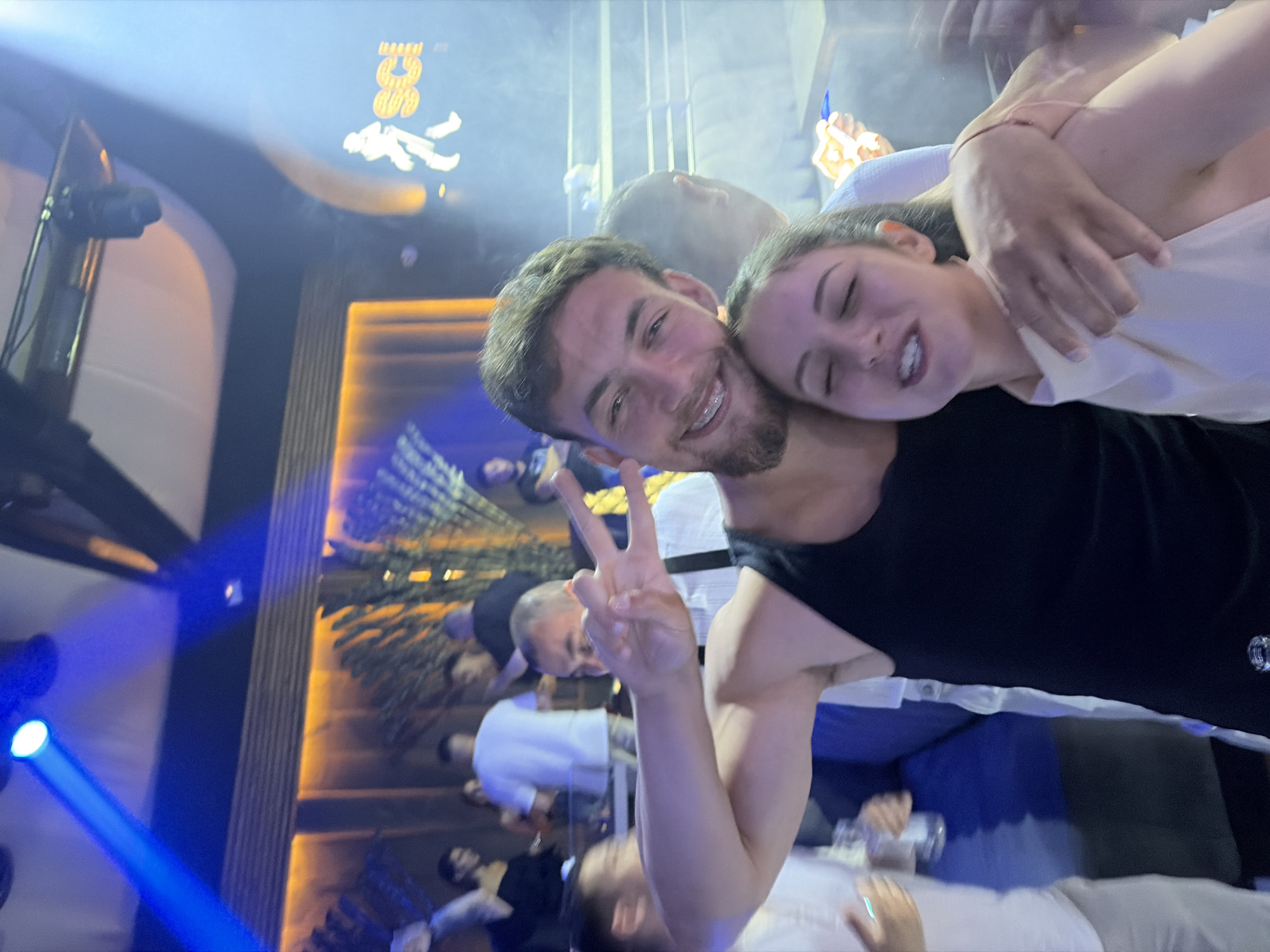 The image depicts an indoor nighttime scene, identified as a club or bar located in Tsarevo, Bulgaria. The environment is characterized by artificial lighting, haze, and numerous individuals socializing.
In the foreground, a young adult male and female are positioned centrally. The male, on the left, has dark hair, a short beard, and is wearing a black tank top. He smiles broadly towards the camera, revealing braces on his upper teeth, and makes a peace sign (V-sign) with his raised right hand. His left arm is around the female. The female, on the right, also appears to be a young adult with dark hair tied back, wearing a light-colored, possibly off-white or pale pink, halter-neck top. Her eyes are closed, and her mouth is slightly open, suggesting an expression of singing or laughter, as she leans her head back against the male.
Behind the primary couple, several other individuals are visible, many blurred due to motion and depth of field. One male directly behind the primary male is also making a peace sign. Patrons are dressed casually, with white t-shirts and dark tops discernible. The setting appears crowded.
The background features distinct architectural and lighting elements. A bright blue spotlight beam originates from the top-left of the frame, cutting across the upper portion of the scene. Warm, orange backlighting illuminates textured wall panels or decorative elements, possibly large artificial plant fronds, further back. On the far right wall, a white illuminated silhouette of a walking figure is displayed next to the yellow letters "SCI", indicating the venue's name, "Sense Club & International". The ceiling displays a modern design with dark beams and lighter panels. A visible haze or smoke is diffused throughout the air, contributing to the club ambiance.