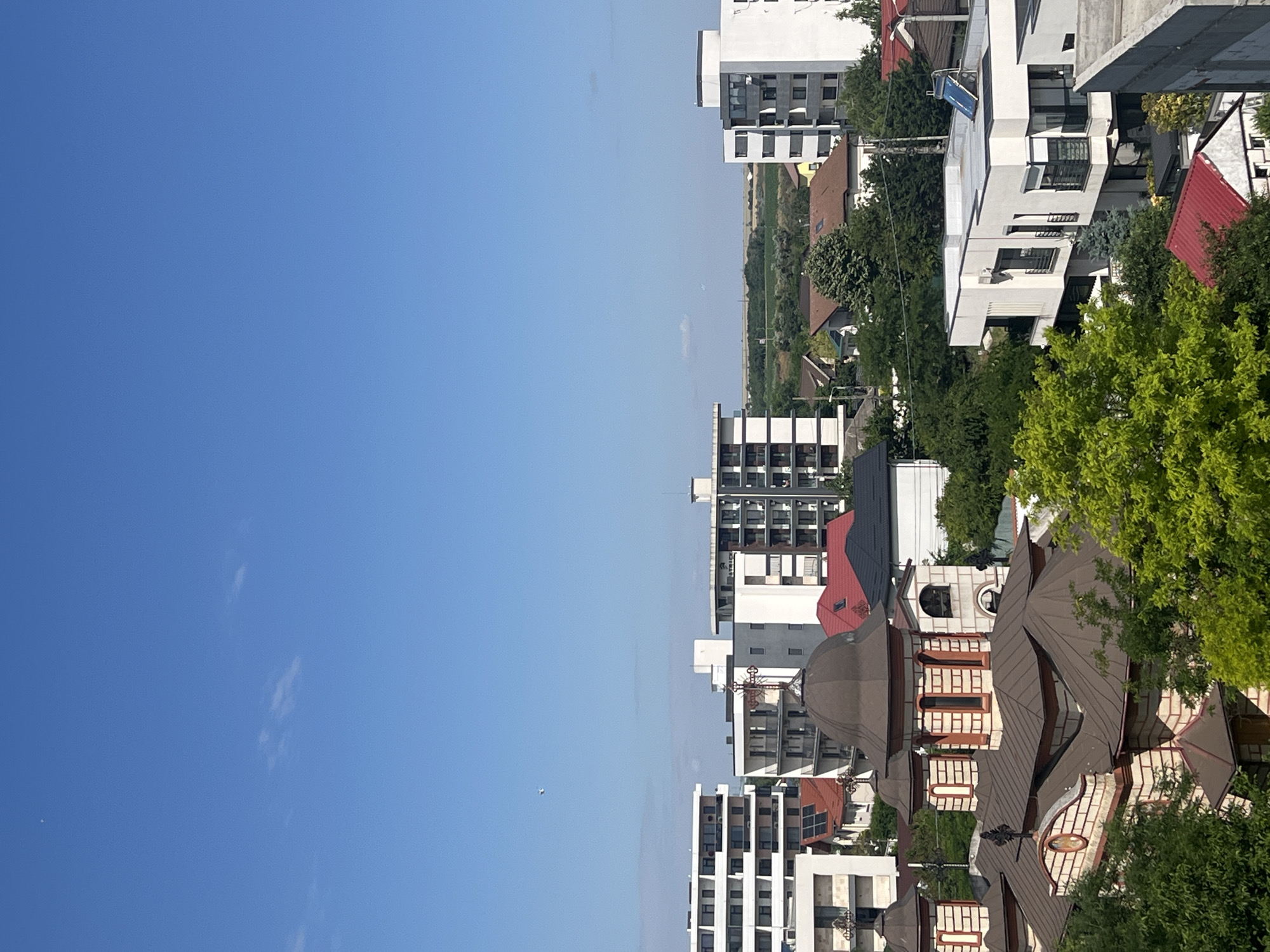 An eye-level, medium shot captures an urban landscape under a clear, bright blue sky in Năvodari, Romania. The scene is dominated by a variety of buildings, ranging from modern apartment blocks with white facades and dark-trimmed windows to a more traditional, ornate church with a brown, domed roof and light brickwork accented with red trim. Several houses with red tile roofs are interspersed among the structures. Lush green trees and foliage are prevalent, creating a contrast with the built environment. In the far distance, a flat, agricultural landscape can be seen. A faint, distant object, possibly an airplane or drone, is visible in the sky. The overall impression is of a sunny day in a mixed residential and urban area.