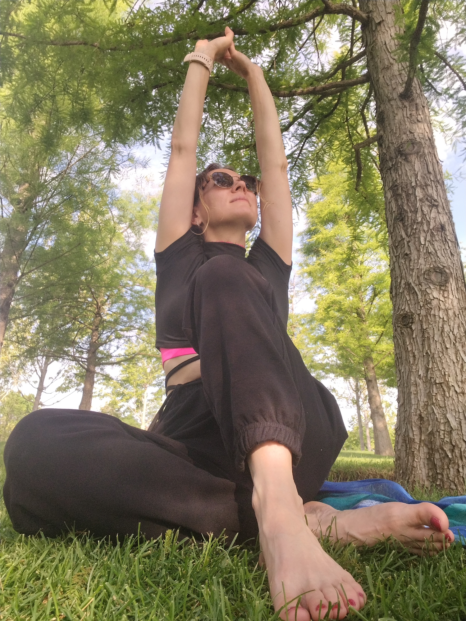 A low-angle outdoor shot shows a woman with blonde hair and sunglasses sitting cross-legged on a grassy lawn. She is wearing a black t-shirt and black pants with a pink sports bra visible underneath. Her arms are raised above her head, and her hands are clasped. She is looking upwards, towards the sky. A tree trunk is prominent to the right of the frame, and other trees and foliage are visible in the background under a bright, possibly sunny sky. A blue and green patterned blanket is partially visible beneath her. The woman's feet are bare and resting on the grass, with red nail polish on her toes. The setting appears to be a park or wooded area within the city of Krasnodar, Russia.