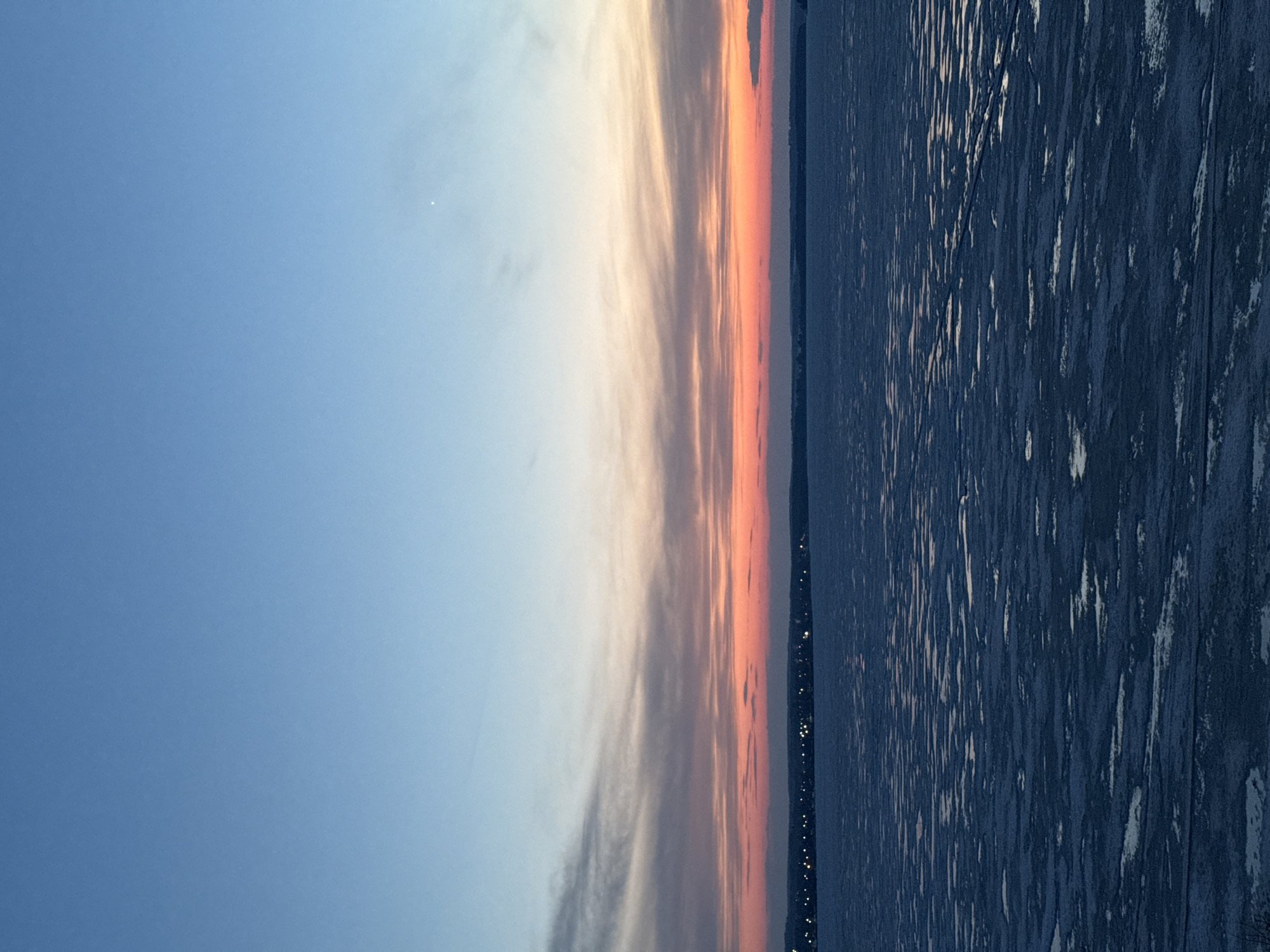 The image depicts a twilight scene in Votkinsk, Russia. A gradient of blue sky transitions to a band of orange and pink clouds near the horizon. The city's lights are visible as small points along the distant shoreline. The foreground shows a dark, textured expanse of frozen water, potentially a lake or river, reflecting some light. The photograph appears to be taken in winter, suggested by the frozen water body. There are no people visible in the image.