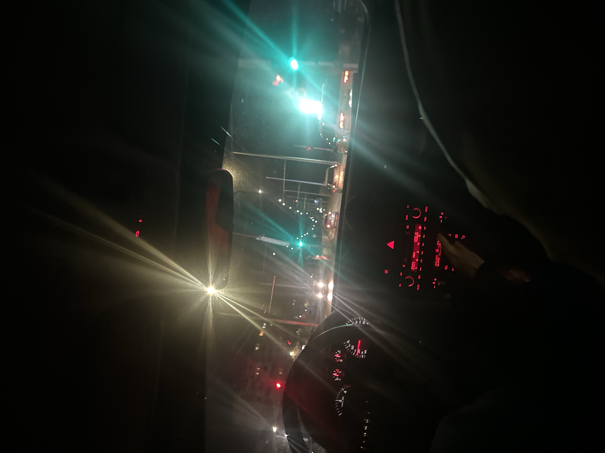 The image depicts the interior of a car at night, likely in Varna, Bulgaria, though this is speculative. The scene is viewed from within the vehicle, focusing on the dashboard and the road visible through the rearview mirror. The dashboard includes the instrument cluster (steering wheel, gauges), a car radio with buttons and a display screen, and interior elements. A hand is visible near the car radio, suggesting someone is interacting with it. The rearview mirror shows a street with traffic lights, and other cars and street lights are visible through the mirror, indicative of a dense city environment at night. The ambient lighting is low with bright lights from the street and traffic lights.