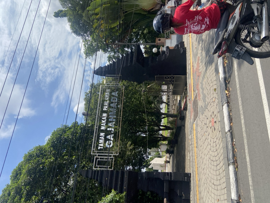 The image shows an entrance to a park in Mojokerto, Indonesia, located at coordinates -7.476, 112.44. The entrance is marked by two large black pillars with an archway between them. Above the archway is a silver sign that reads “Taman Makam Pahlawan Gajahmada.” The sign is situated in a frame of iron bars. The park appears to be a cemetery or memorial area with a green lawn and trees visible beyond the entrance. A person in a red shirt and a helmet is riding a black motorcycle toward the entrance. The motorcycle is stopped in the road just outside the park’s entrance. The ground in the foreground is a paved road with a cobblestone walkway leading to the entrance. The road is lined with trees, and the sky is a clear blue with white clouds. The scene is bathed in bright sunlight.