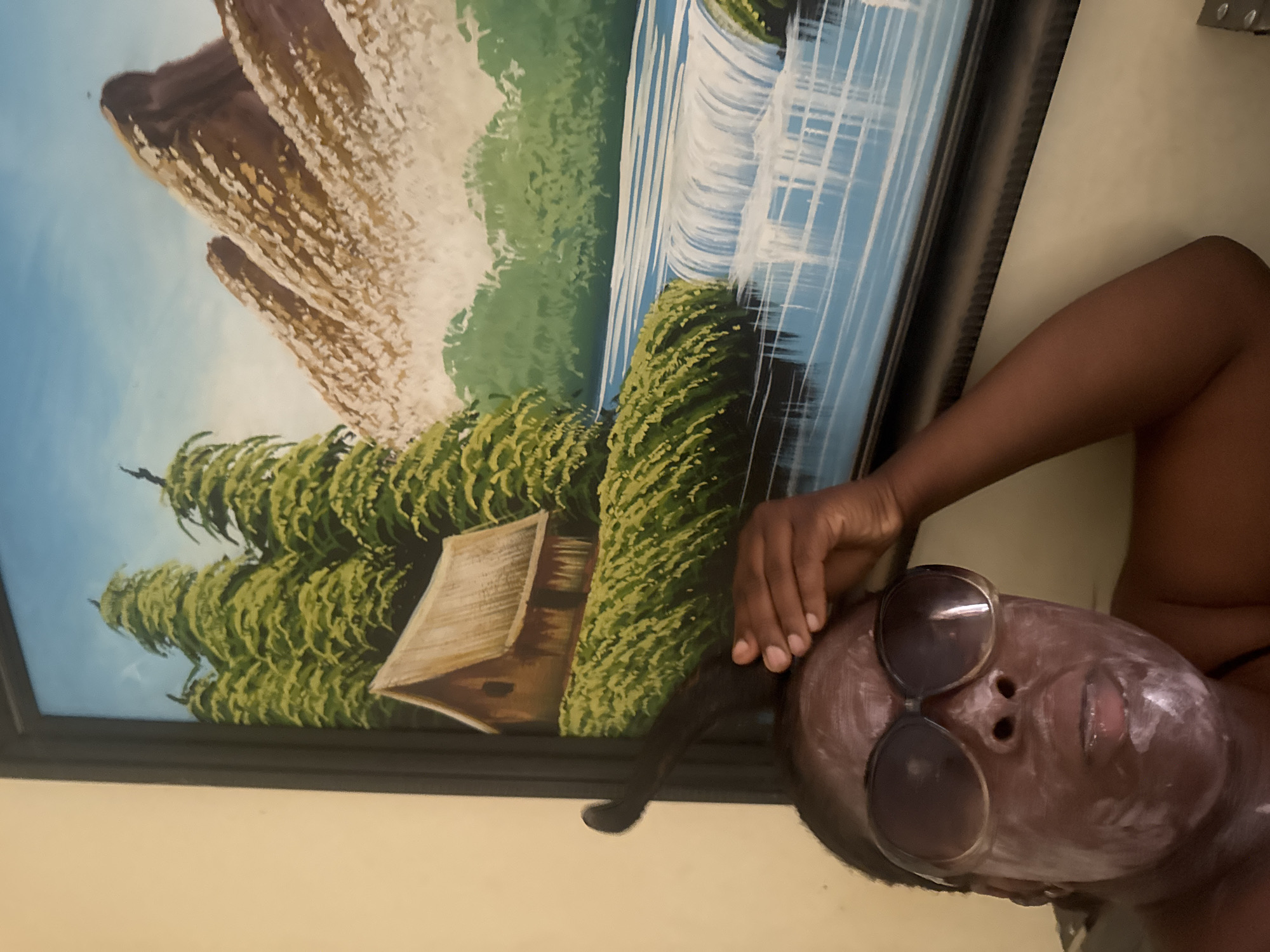 A person is lying on a light-colored surface, with an arm raised towards a painting hanging on a pale yellow wall. The person has sunglasses perched on their face, which is covered in a white substance, possibly a face mask. The painting, framed in black, depicts a landscape scene with what appears to be a waterfall, green vegetation, and rocky formations. The location is New Karu, Nigeria.
