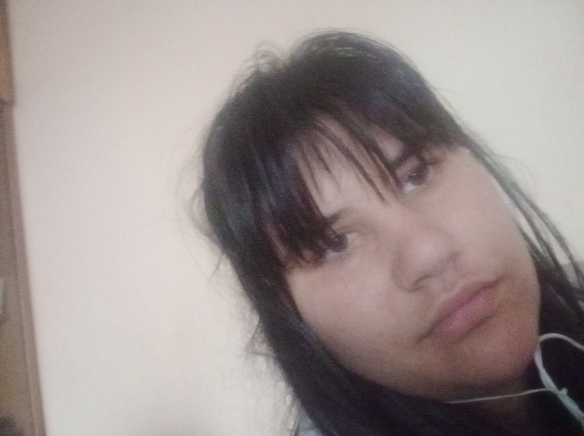 The image is a close-up, slightly tilted portrait of an individual from the chest up. The person has dark, straight hair with bangs that partially cover the forehead. The face is directed towards the camera, displaying dark eyes, a nose, and closed lips with a neutral expression. A white earbud cable is visible across the lower right portion of the frame, resting against dark clothing worn by the individual. The background consists of a plain, light-colored wall, possibly beige or off-white, indicating an indoor setting. A dark, vertical object, consistent with a wooden frame or furniture edge, is partially visible on the far left edge of the image. The image quality shows a noticeable grain. The lighting appears soft and diffused. This scene is located within an indoor environment in Cherven Bryag, Bulgaria.
