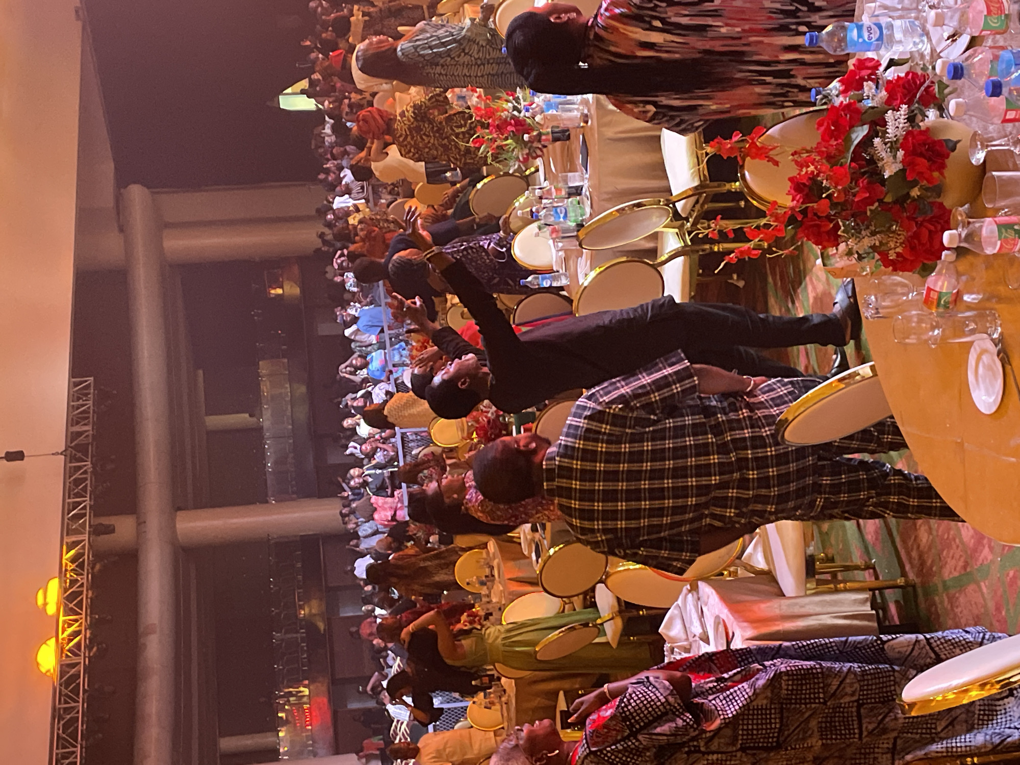 The image shows a large indoor event in Itirin, Nigeria. A large crowd is seated at tables covered with white tablecloths. Many people are in formal attire. Red flower arrangements and water bottles adorn the tables. Some people are gesturing upwards. A multi-tiered balcony is visible along the wall, with more attendees viewing from above. The event appears to be taking place in a large hall with structural support beams. Lighting fixtures are attached to a metal structure on the left.