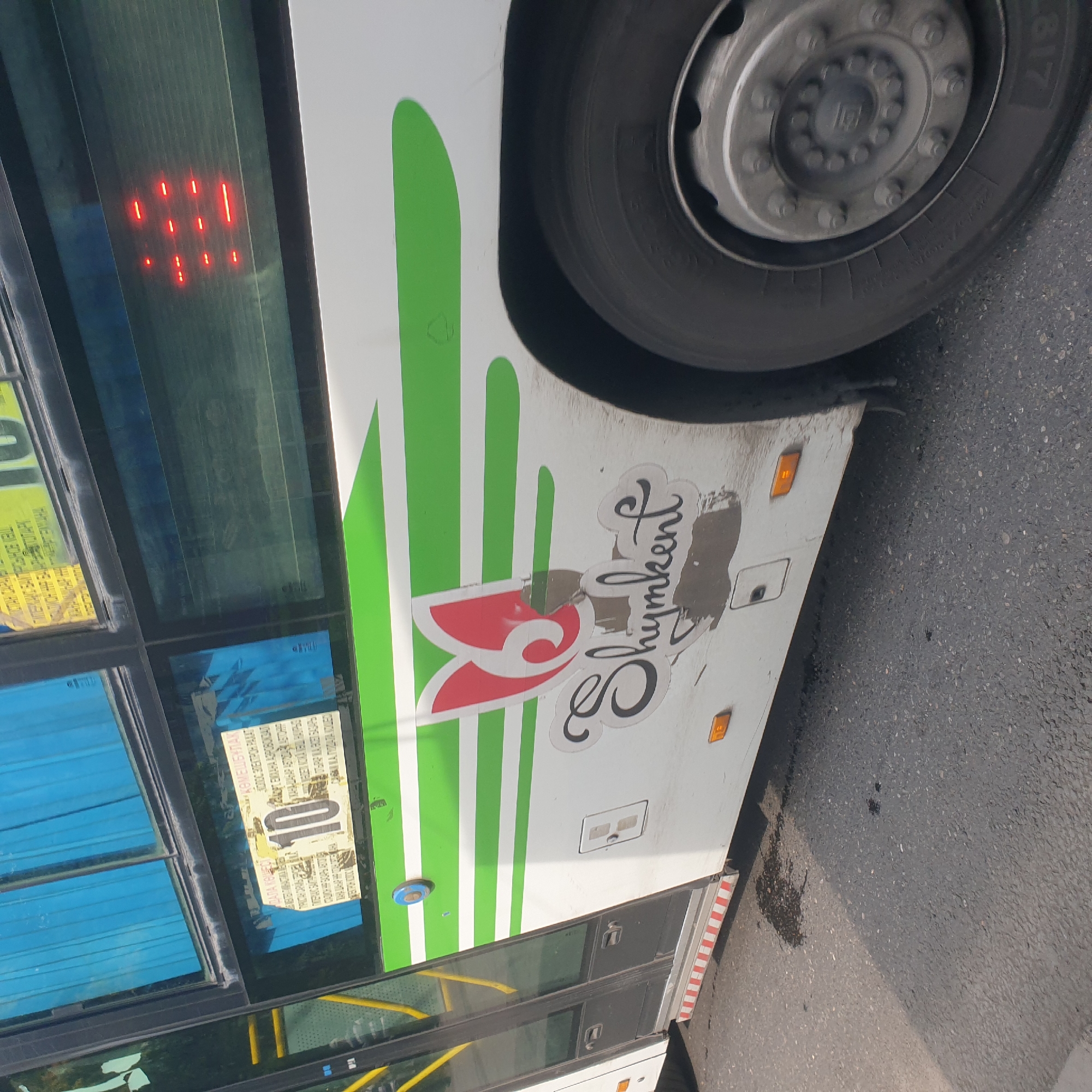 The image shows the side of a white bus with green stripes and a stylized red logo on its side that says "Shymkent." There is a sticker on the window with the number "10" in a circle and some text in what appears to be Kazakh or Russian. The bus is parked on a street with asphalt pavement. There are some dark stains on the pavement near the bus. The overall scene is of a bus in an urban environment, likely in the city of Shymkent, Kazakhstan, given the text on the bus.