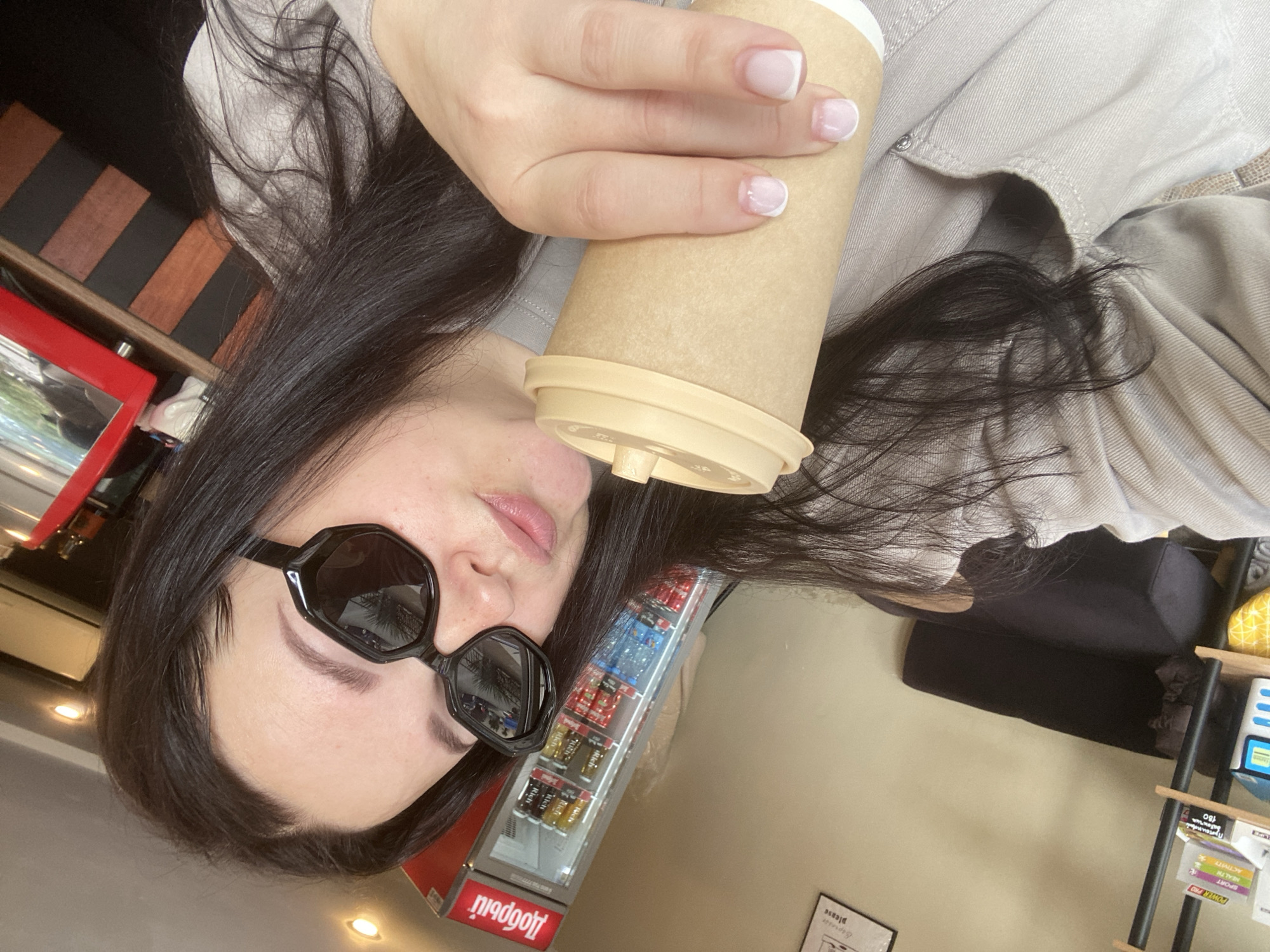 A young woman with dark hair, wearing black sunglasses, is holding a coffee cup in her right hand. She is indoors, likely in a cafe or store. Behind her, there's a refrigerated display case filled with various canned or bottled drinks. A sign on the display case seems to have Russian text. To her left, there is dark wood paneling and a red framed object. To her right, there appears to be a shelf with books and some other items. The woman is wearing a light-colored shirt or jacket. The location appears to be Sochi, Russia based on text visible on the display case.