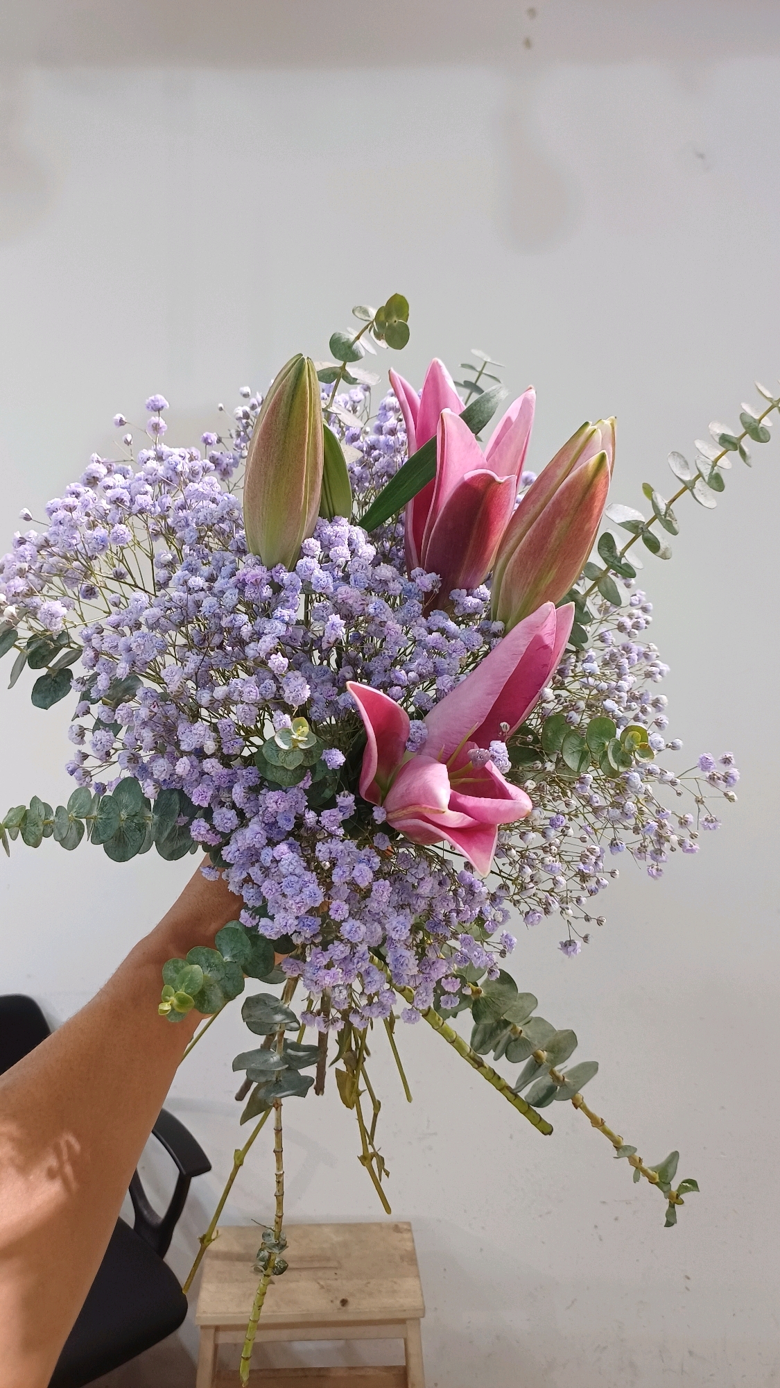 A hand holds a bouquet of pink lilies and small purple flowers, likely baby's breath, with eucalyptus sprigs, against a plain white wall. The bouquet is the primary subject, with a focus on the vibrant colors and delicate textures of the flowers. The background is minimally detailed, suggesting an indoor setting or a clear outdoor space. No specific geographical markers are visible that would place this scene in Kuala Lumpur, Malaysia.