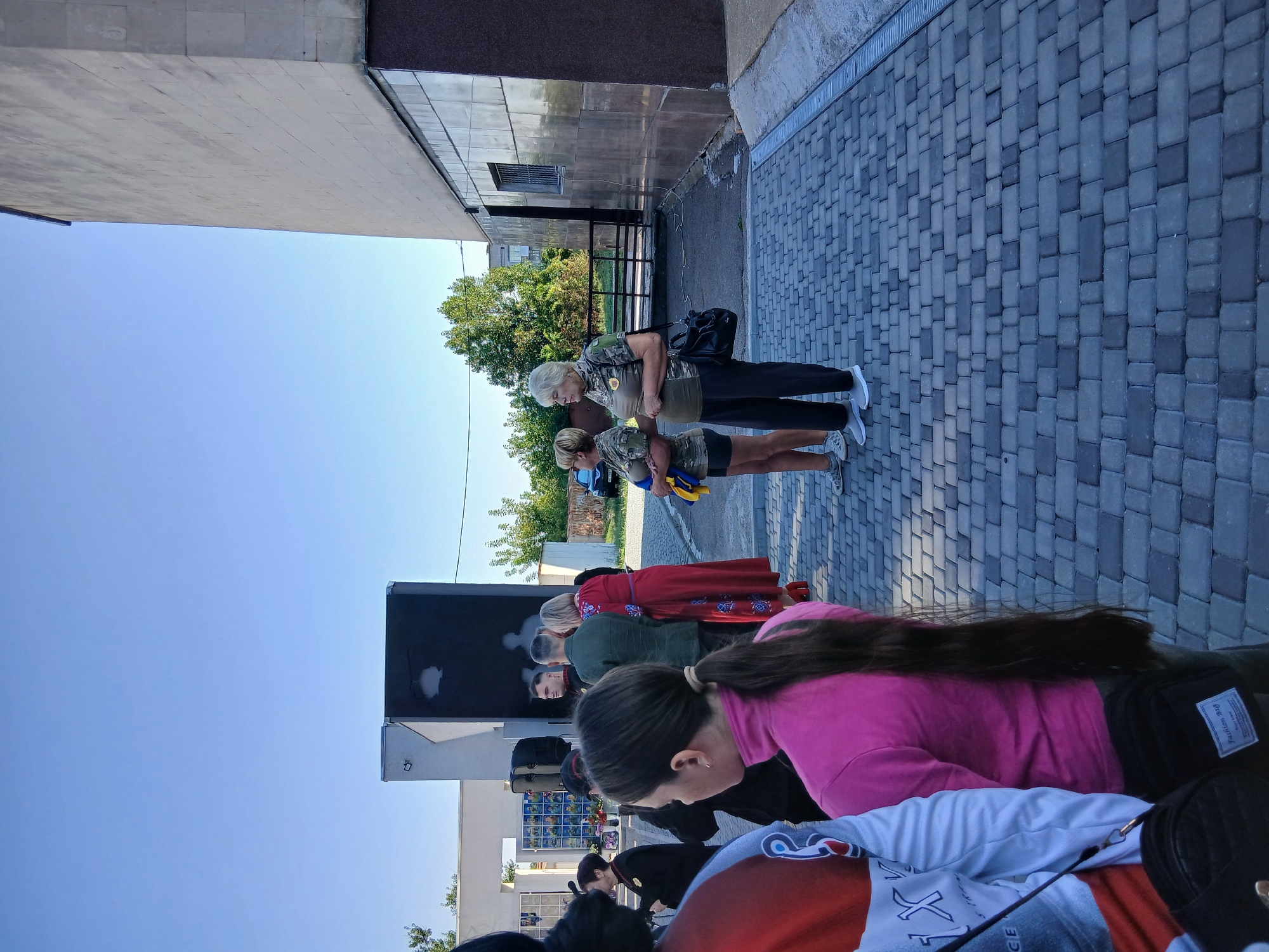 The image shows a group of people standing outside on a paved area in front of a building. The building is modern, with a light-colored facade and dark accents. There are trees and a clear blue sky in the background. The people appear to be gathered for an event, possibly a ceremony or commemoration, as some are wearing military-style uniforms and holding Ukrainian flags. One woman in the foreground is wearing a bright pink top and has long dark hair. Another woman in the background is wearing a red dress. The paved area is made of interlocking bricks. The scene appears to be taking place in the city of Verkhnodniprovsk, Ukraine.