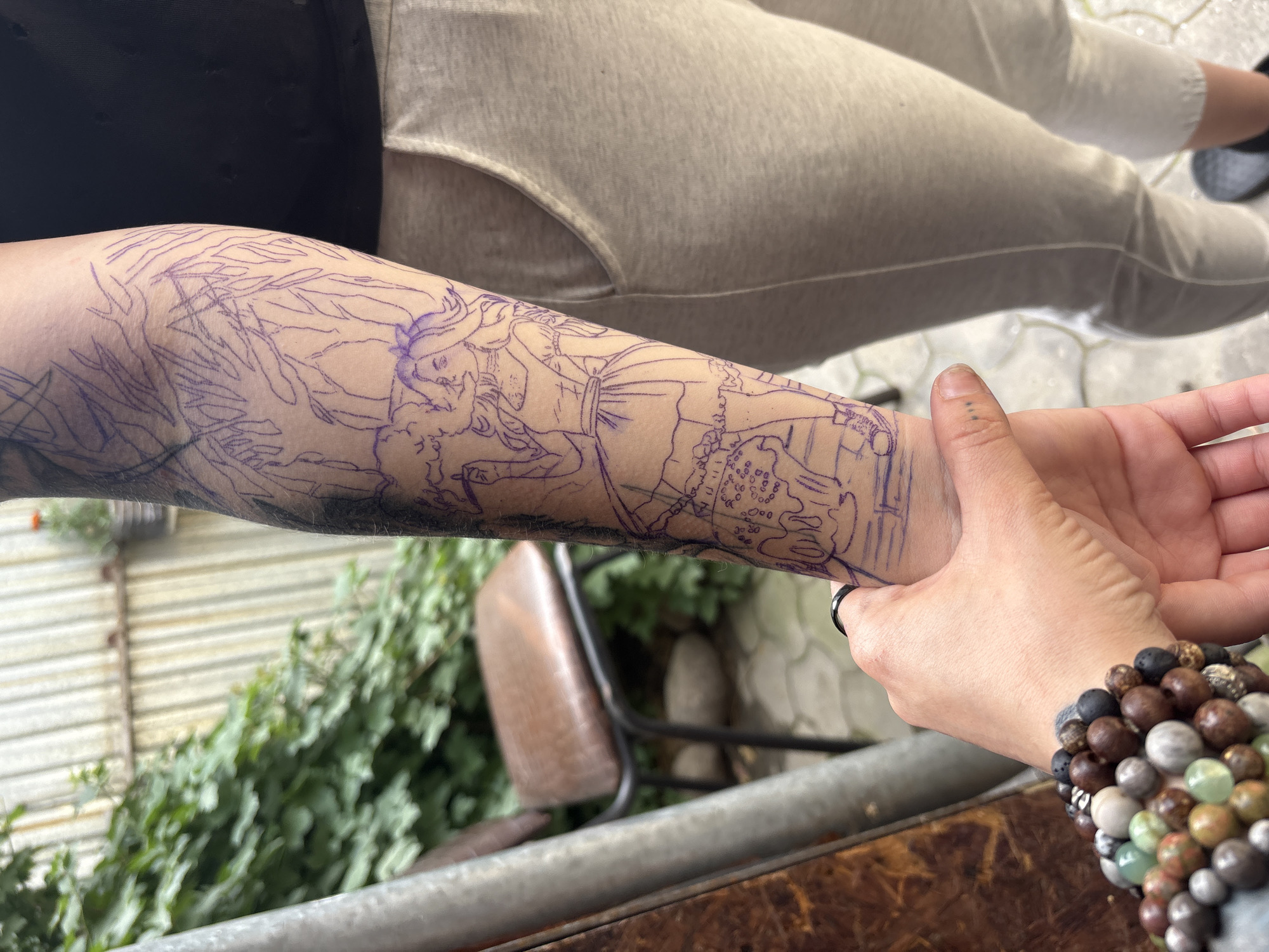 An individual's forearm, marked with a purple tattoo outline of a woman in a dress and trees, is prominently displayed. The tattoo design features a woman with flowing hair holding what appears to be a cigarette, and elements of foliage above her. The forearm is held in the foreground, with a hand adorned with beaded bracelets and a ring visible at the bottom left. In the background, a brown chair with a metal frame sits amidst lush green foliage. A corrugated metal fence forms the backdrop. The ground appears to be paved with light-colored stones, possibly in an outdoor setting. The image does not contain information that would allow for a specific location within Almaty, Kazakhstan.