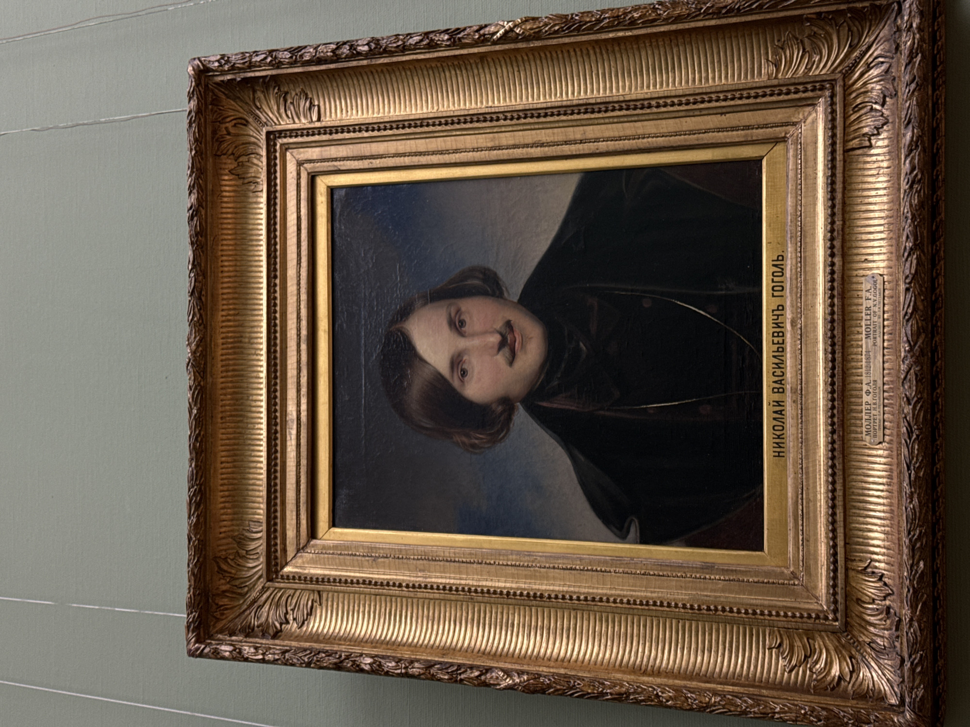 A medium shot captures a framed portrait hanging against a muted green wall. The portrait depicts a man, likely from the 19th century, dressed in dark attire. A label on the frame includes the text "НИКОЛАЙ ВАСИЛЬЕВИЧ ГОГОЛЬ" and "МОЛЛЕР Ф.А. ПОРТРЕТ Н.В.ГОГОЛЯ". Given the subject's identity and the Cyrillic text, the location is most likely within Moscow, Russia, possibly in a museum or gallery.