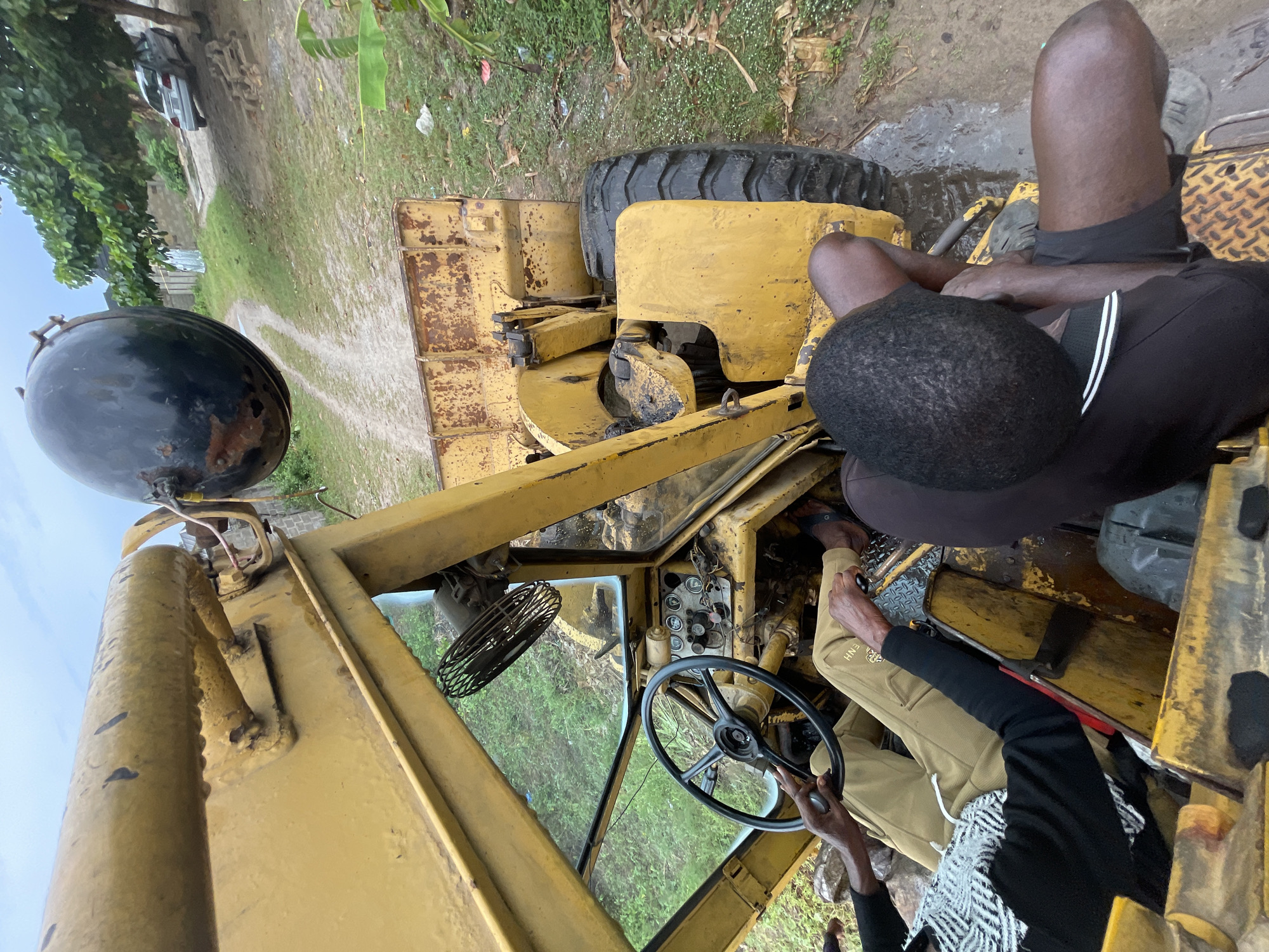 The image depicts a man in the driver's seat of a yellow bulldozer in Badagry, Nigeria. Another man sits on the bulldozer's hood. The environment appears to be a grassy area, with a dirt road in the background. A vehicle is also visible in the background. The bulldozer is old and shows signs of wear and tear.