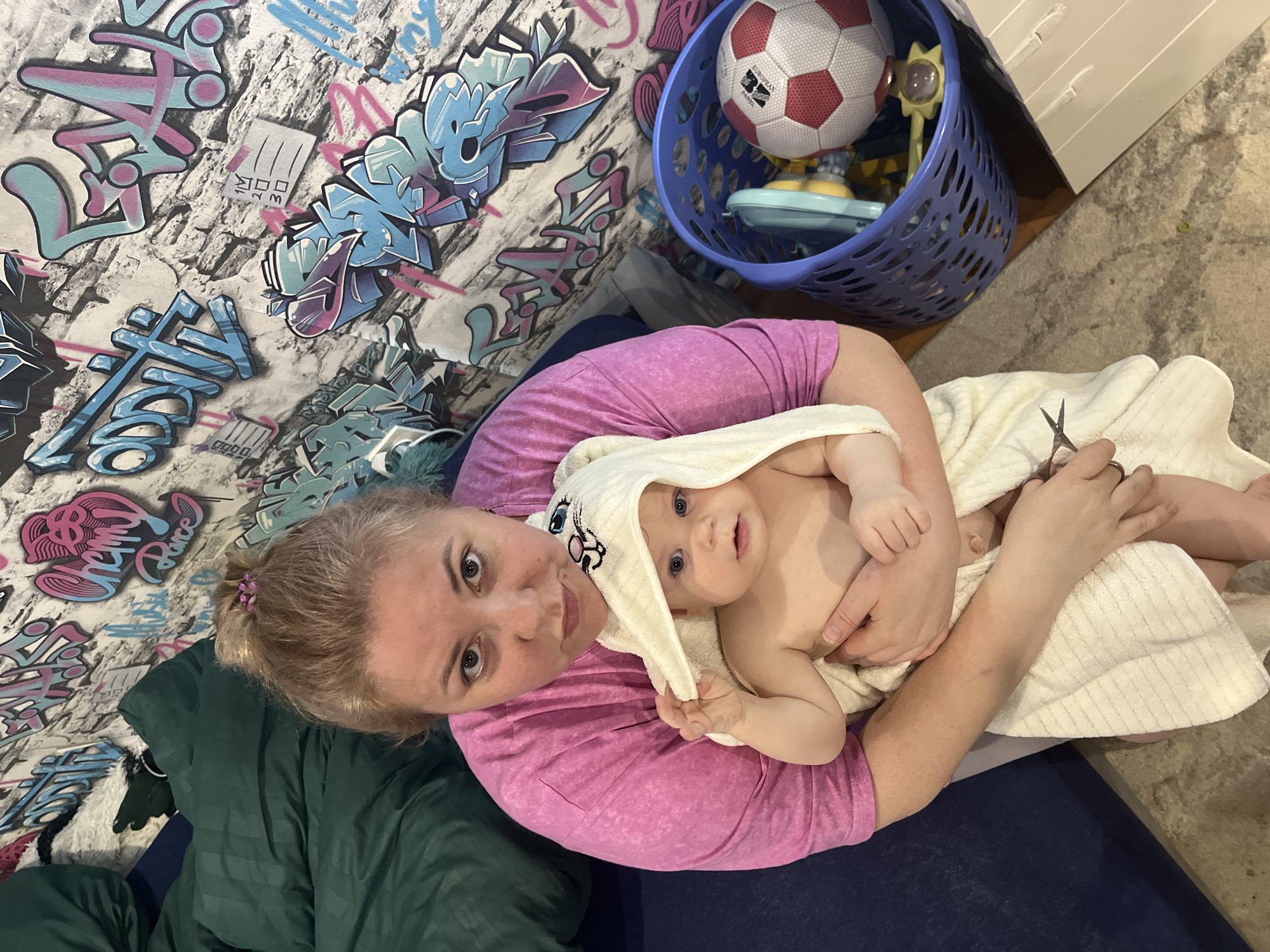 A woman wearing a pink shirt and a blonde ponytail is holding a baby wrapped in a white hooded towel. The woman is looking at the camera while holding scissors. The baby is also looking at the camera. To the left of the woman is a dark green blanket and a wall with graffiti. A blue basket with toys and a soccer ball is to the right and behind the woman. The location appears to be an indoor residence, but the city of Vesyoloye, Russia, cannot be confirmed based on the provided visual information.