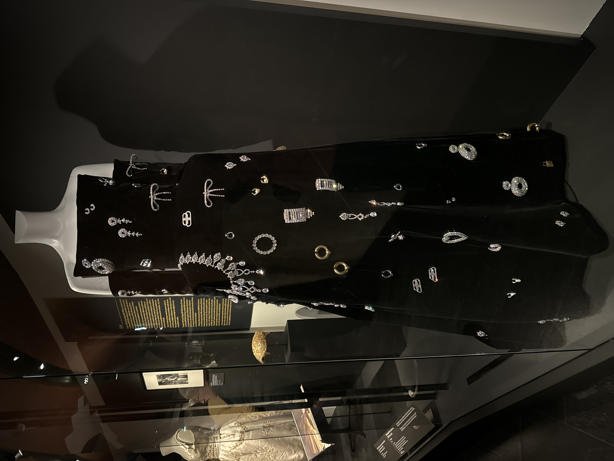 A black dress embellished with various jewelry pieces is displayed on a mannequin in a glass case, presumably in a museum exhibit. The jewelry includes diamonds, gold pieces, and a rhinestone-like crown. The display includes information panels and is likely located in Paris, France, based on the style of the objects. The exhibit appears to showcase a collection of fashion and jewelry. The surroundings are dimly lit with black backgrounds, and there are reflections on the glass surface of the display case. Other items are displayed in the case below the dress.