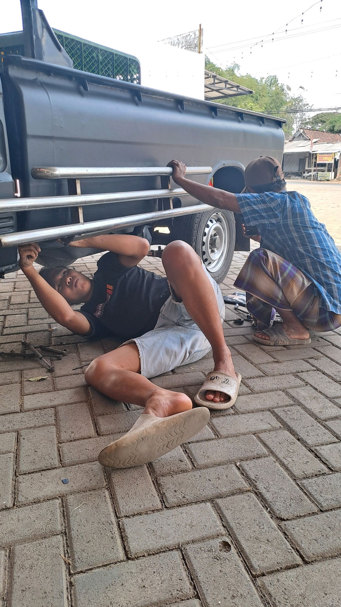 In Kenduruan, Indonesia, two individuals are working on the underside of a black pickup truck. One person, wearing a blue checkered shirt, a brown cap, and checkered pants, is kneeling and appears to be holding onto the truck's frame. The other person, dressed in a black t-shirt and light-colored shorts, is lying on the ground, reaching up to work on the truck. Tools are scattered on the paved ground around them. The scene takes place outdoors, with buildings and trees visible in the background.