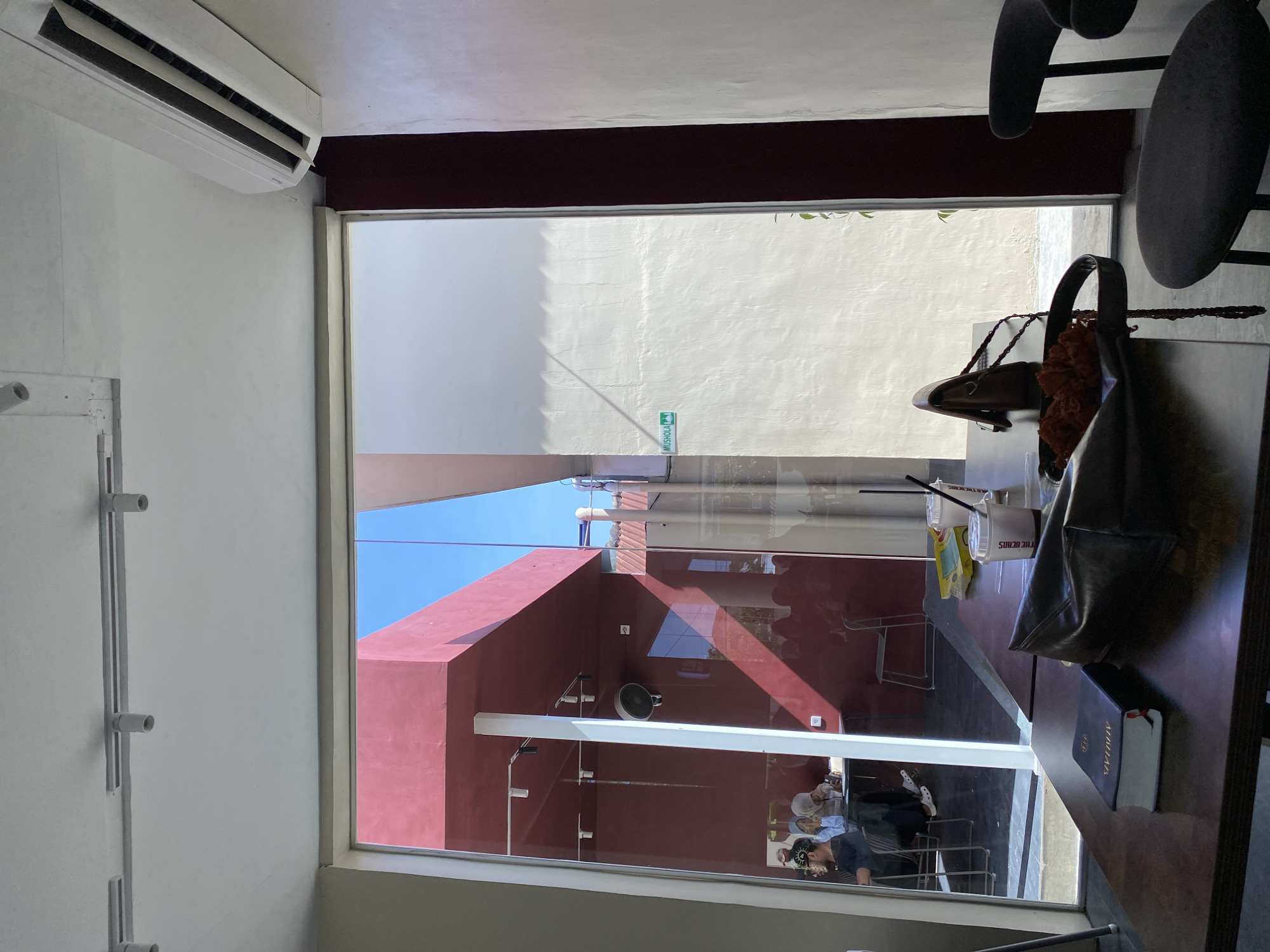 The image shows an interior view through a large window in Lhokseumawe, Indonesia. Outside the window is a street with a red building and a white wall. A street sign is visible, although the text is unclear. Inside, a table occupies the foreground, cluttered with items including a handbag, drinks in plastic cups with straws, a book titled "Alhilal," and other assorted items. Several people are visible through the window, seated in what appears to be an outdoor seating area of the establishment. An air conditioning unit is installed high on the interior wall. A partial view of chairs is present.