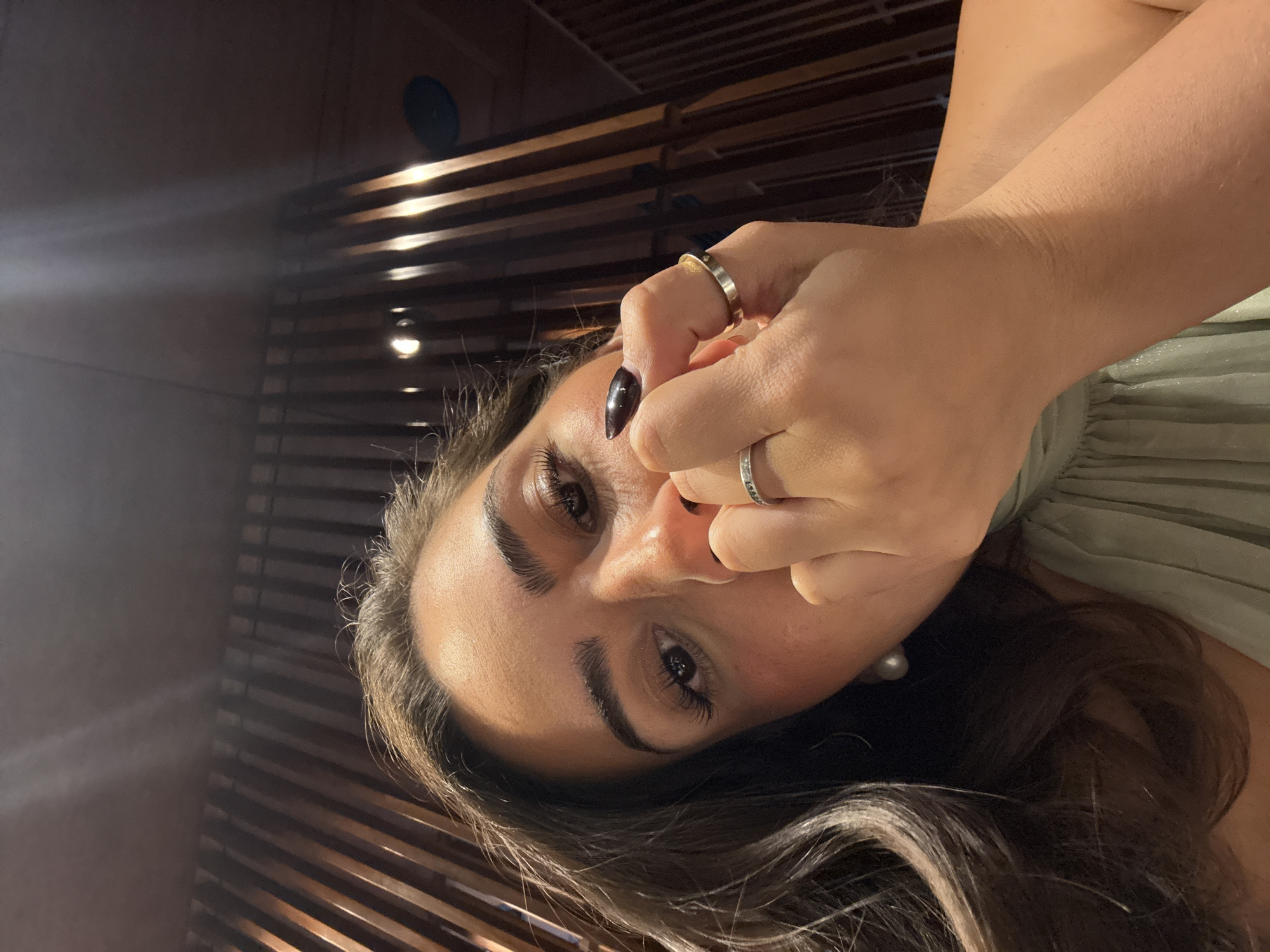 A close-up, rotated portrait captures a woman looking directly at the camera, her head tilted towards the bottom right of the frame. She has long, dark brown hair with subtle lighter highlights, framing her face. Her eyes are open and directly engaging with the viewer, complemented by dark, defined eyebrows and long eyelashes.
Her right hand is positioned near her face, with fingers resting around her nose and chin area. She wears two silver-toned rings: a wider band on her index finger and a thinner one on her ring finger. Her nails are painted a dark, glossy color. A light green or sage-colored garment with soft pleats is visible on her right side, and a single pearl earring adorns her right earlobe.
The background consists of dark, horizontal wooden slats or beams, illuminated by artificial light sources that create warm reflections. This suggests an indoor setting, possibly a restaurant, bar, or lounge. A circular dark object, likely a vent or speaker, is visible in the upper background. The lighting is dim, indicating an evening or night-time ambiance within an establishment in Puebla, Mexico. No visible text is present.