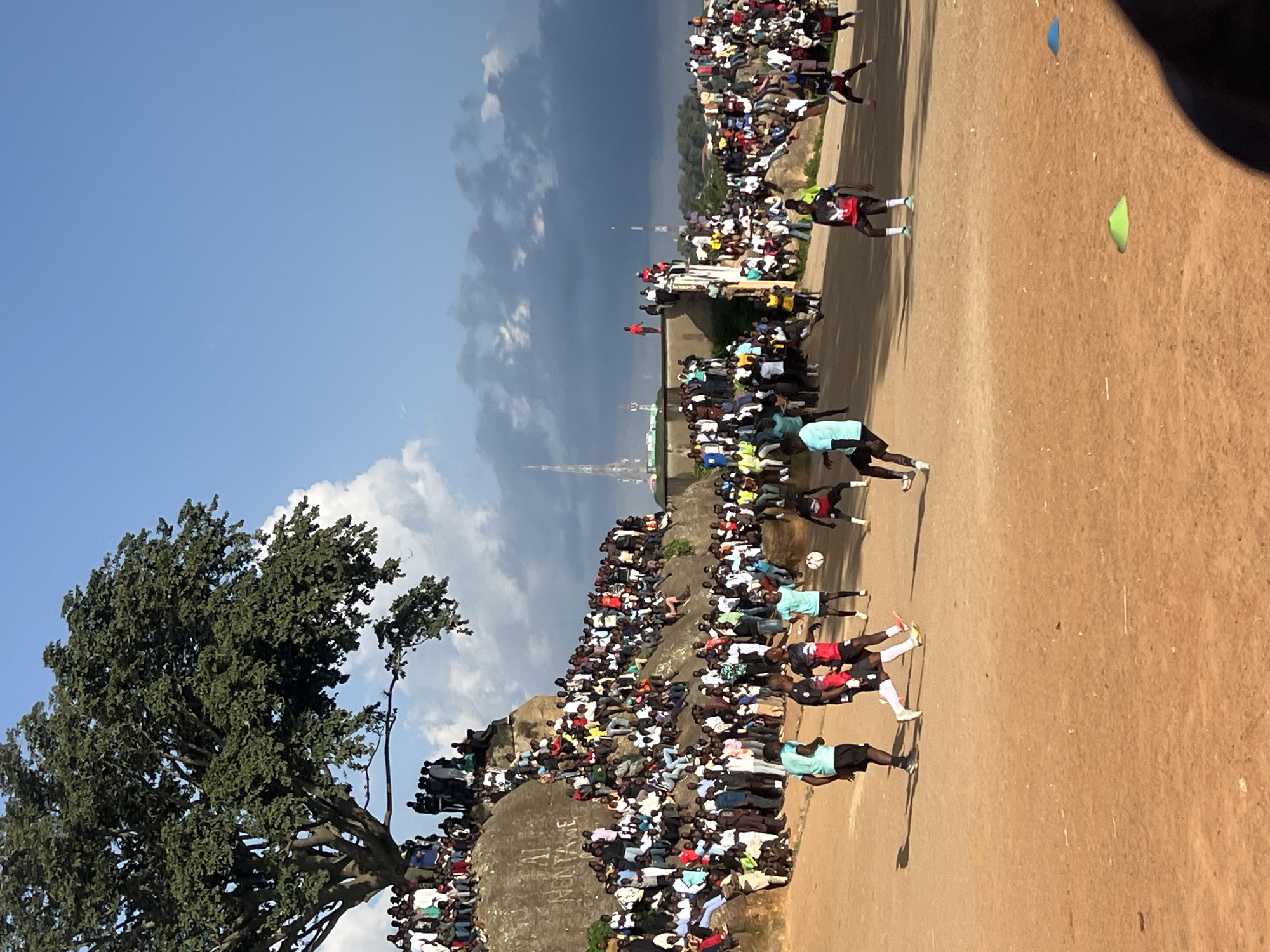 This is a wide shot of a soccer game taking place in a dusty, open field. A large crowd of people, estimated to be hundreds, are gathered on a rocky, elevated area on the left side of the frame, watching the game. Some people are sitting and others are standing. A large, leafy tree is prominent on the top left of the frame, shading a portion of the crowd. A few players in athletic wear are actively participating in the game on the field. In the background, there are buildings, communication towers, and a slightly overcast sky with some darker clouds. The location is identified as Jos, Nigeria, specifically at the "Rayfield Rock" area known for its natural rock formations and its use as a public gathering space.